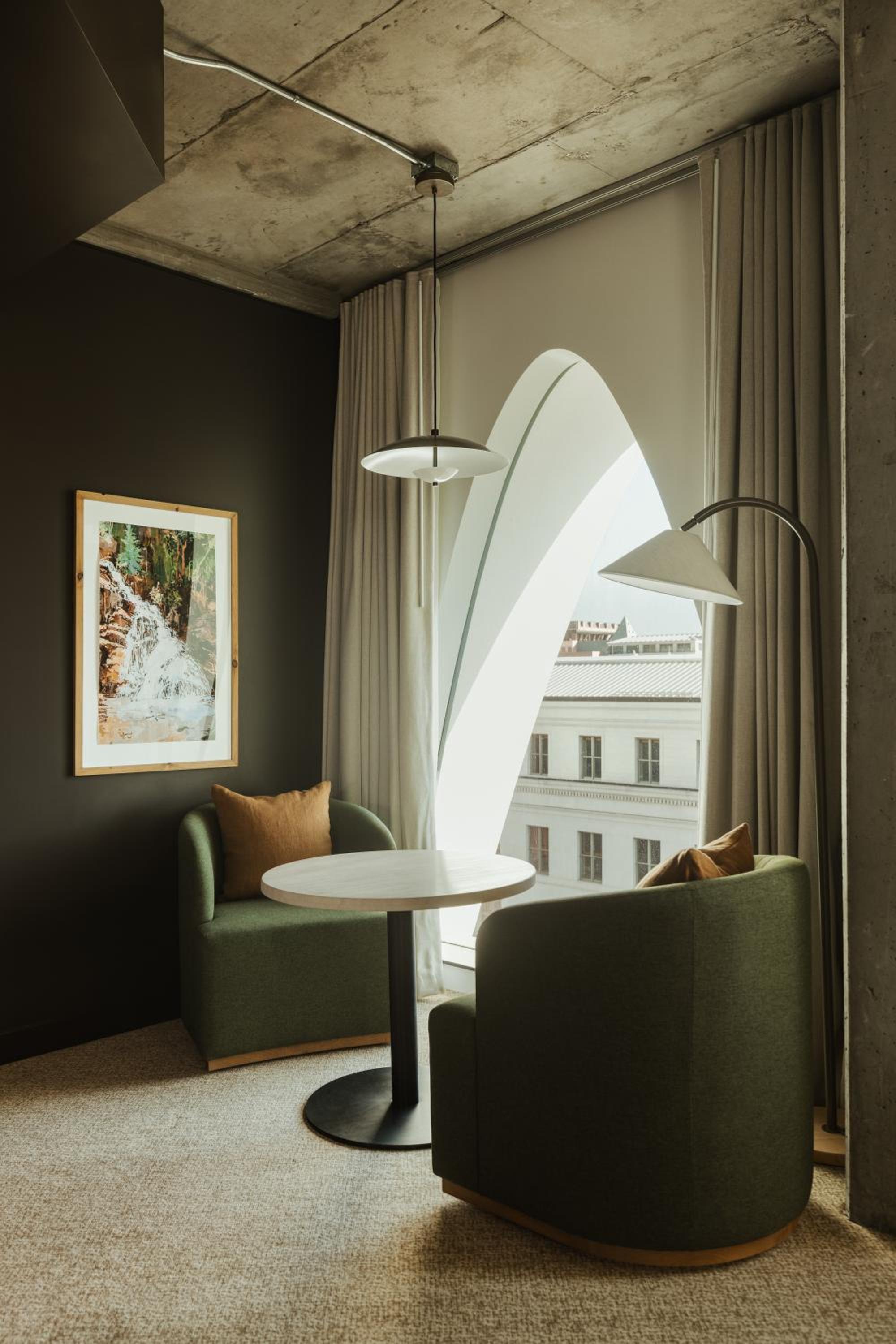 Green armchairs and a round table set beside a tall arched window in a corner nook.