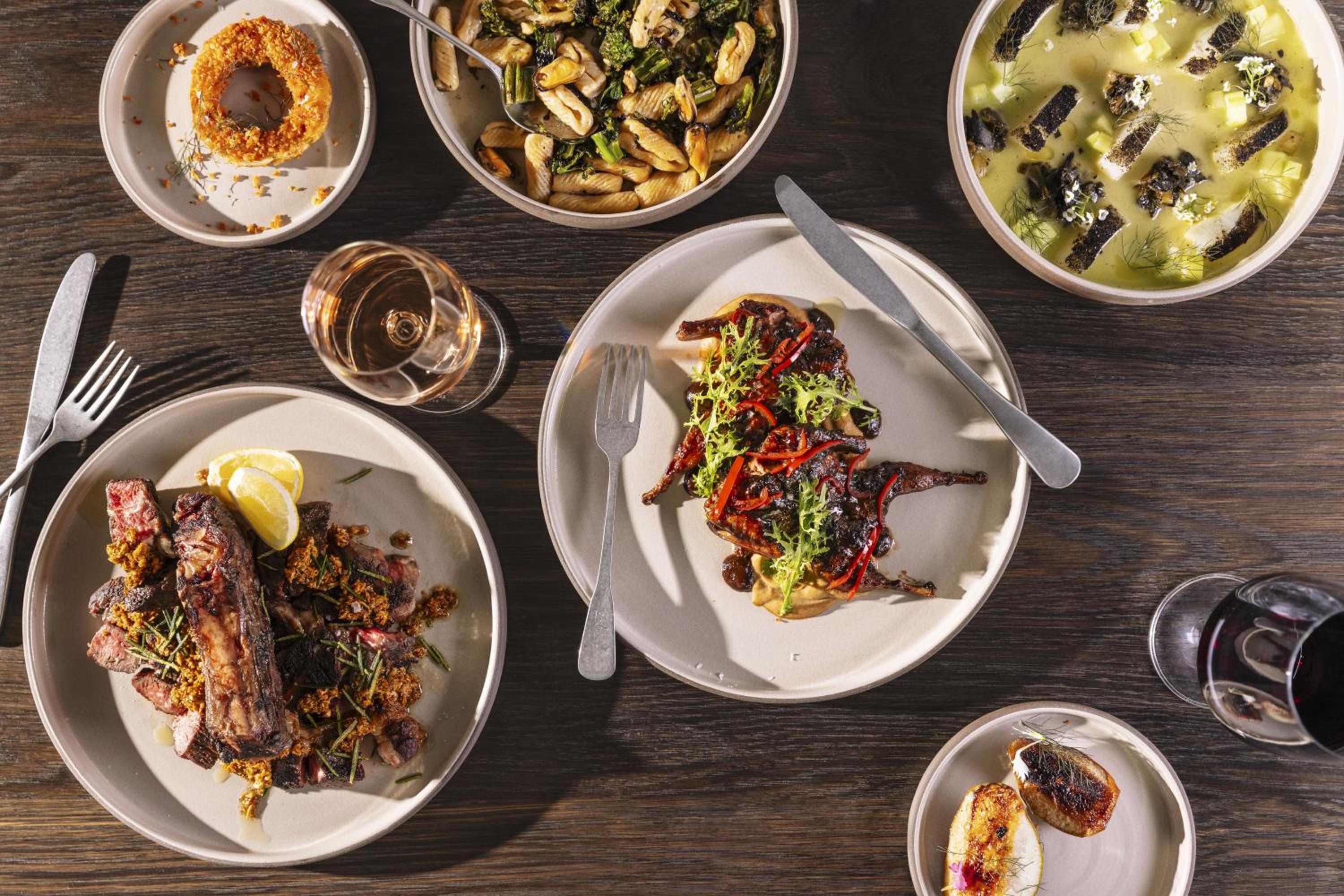 Overhead view of dinner plates with roasted meats, pasta, and vegetable dishes spread across a dark wood table.