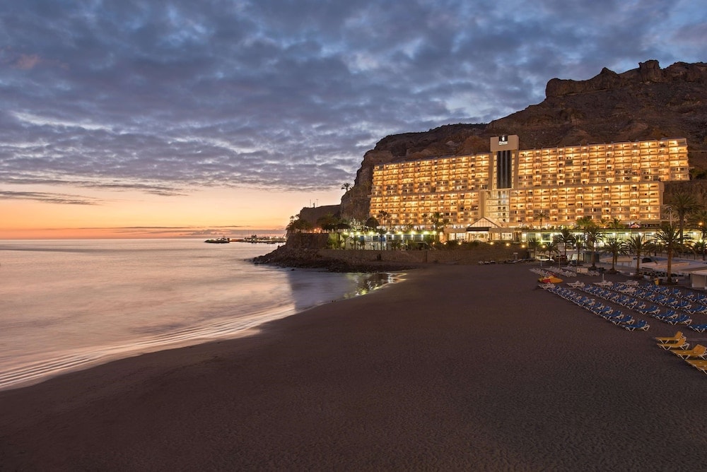 Illuminated hotel facade of Princess Taurito set against a rocky cliff beside the beach at sunset.