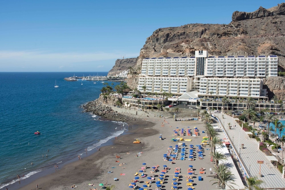 Elevated view of a dark sand beach with sun loungers fronting the resort against rugged cliffs and the sea.