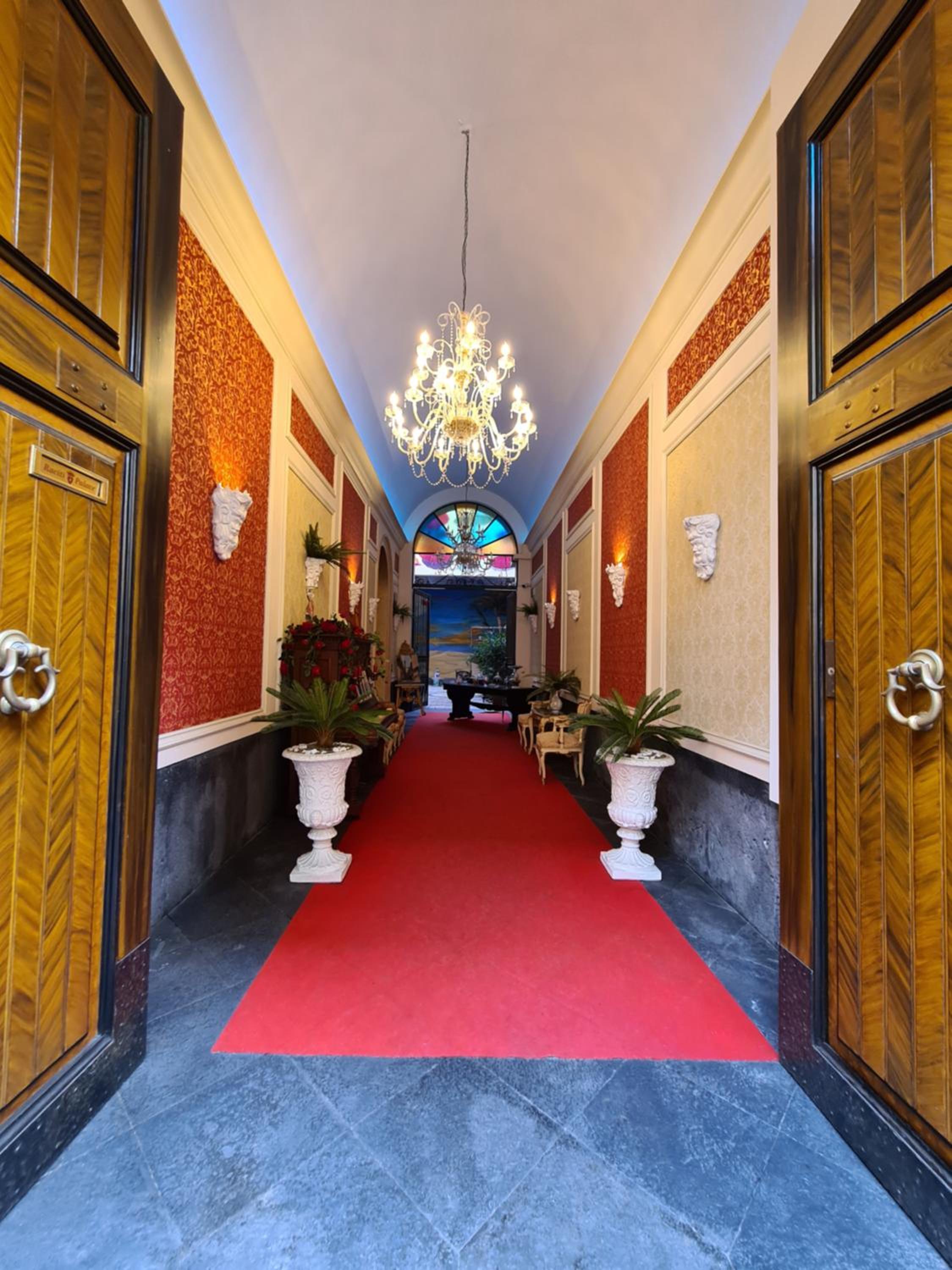 Hallway at Raciti Palace lined with red damask wallpaper and a red runner beneath a crystal chandelier.