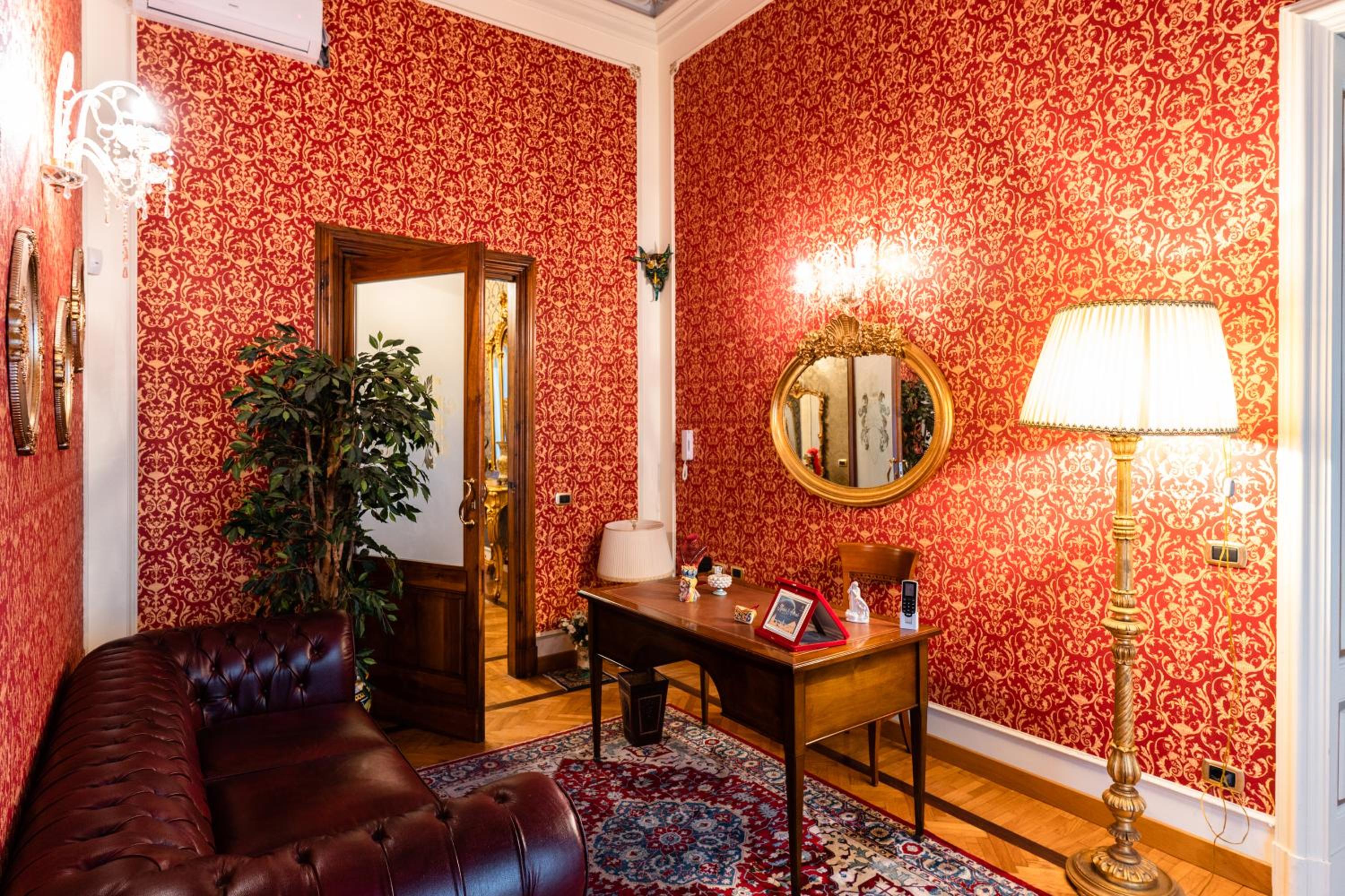 A room with red and gold damask wallpaper, a leather tufted sofa, a wooden desk, and a standing floor lamp.