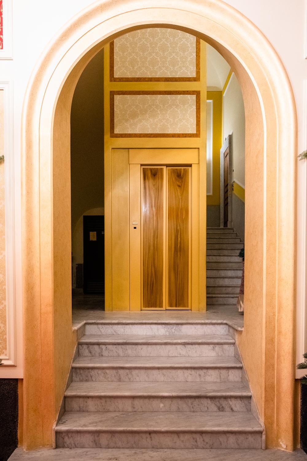 Marble steps viewed through an archway, leading to an elevator with wooden doors and an adjoining staircase.