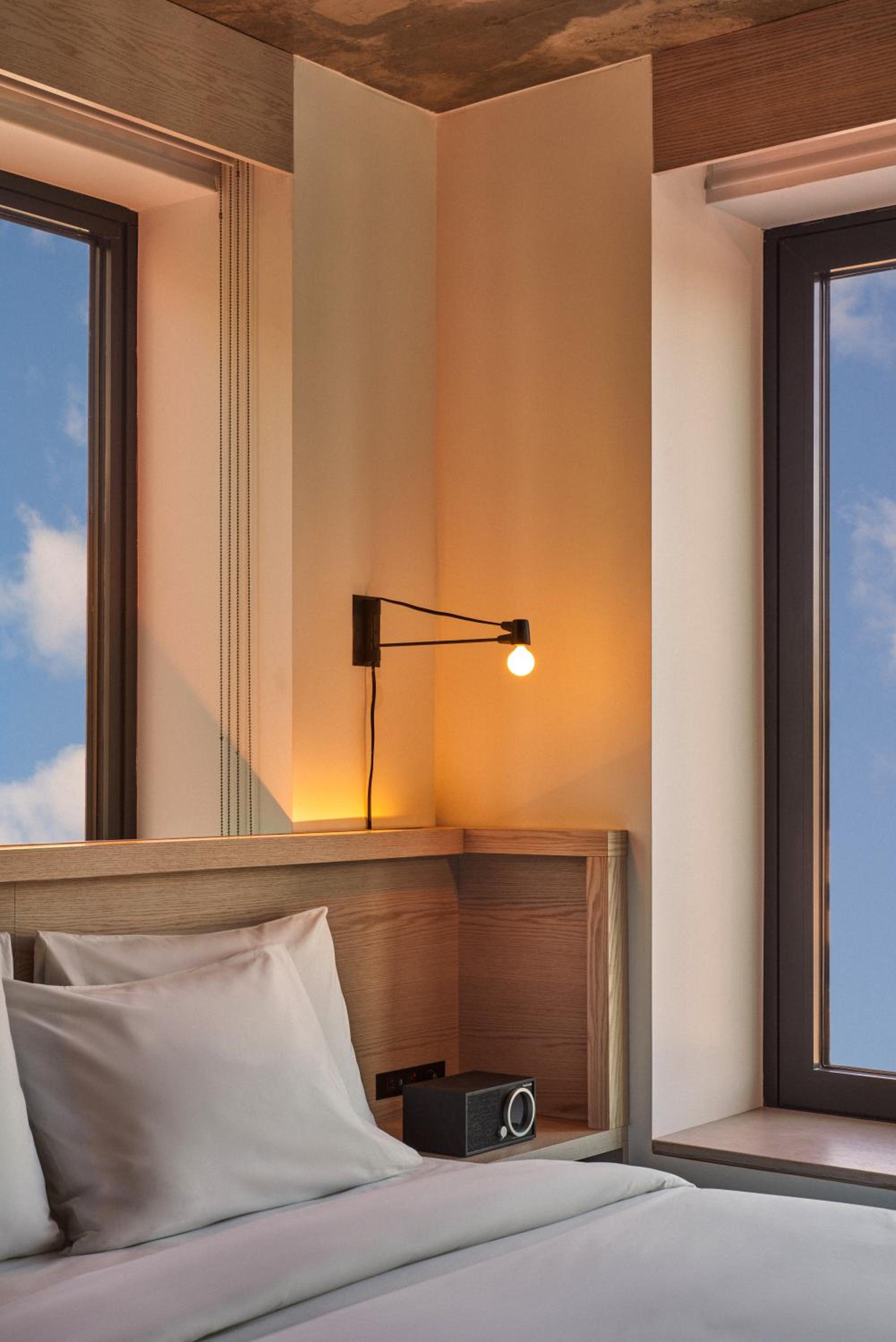 Hotel bedroom corner with a light wood headboard, bedside reading lamp, and windows framing a blue sky.