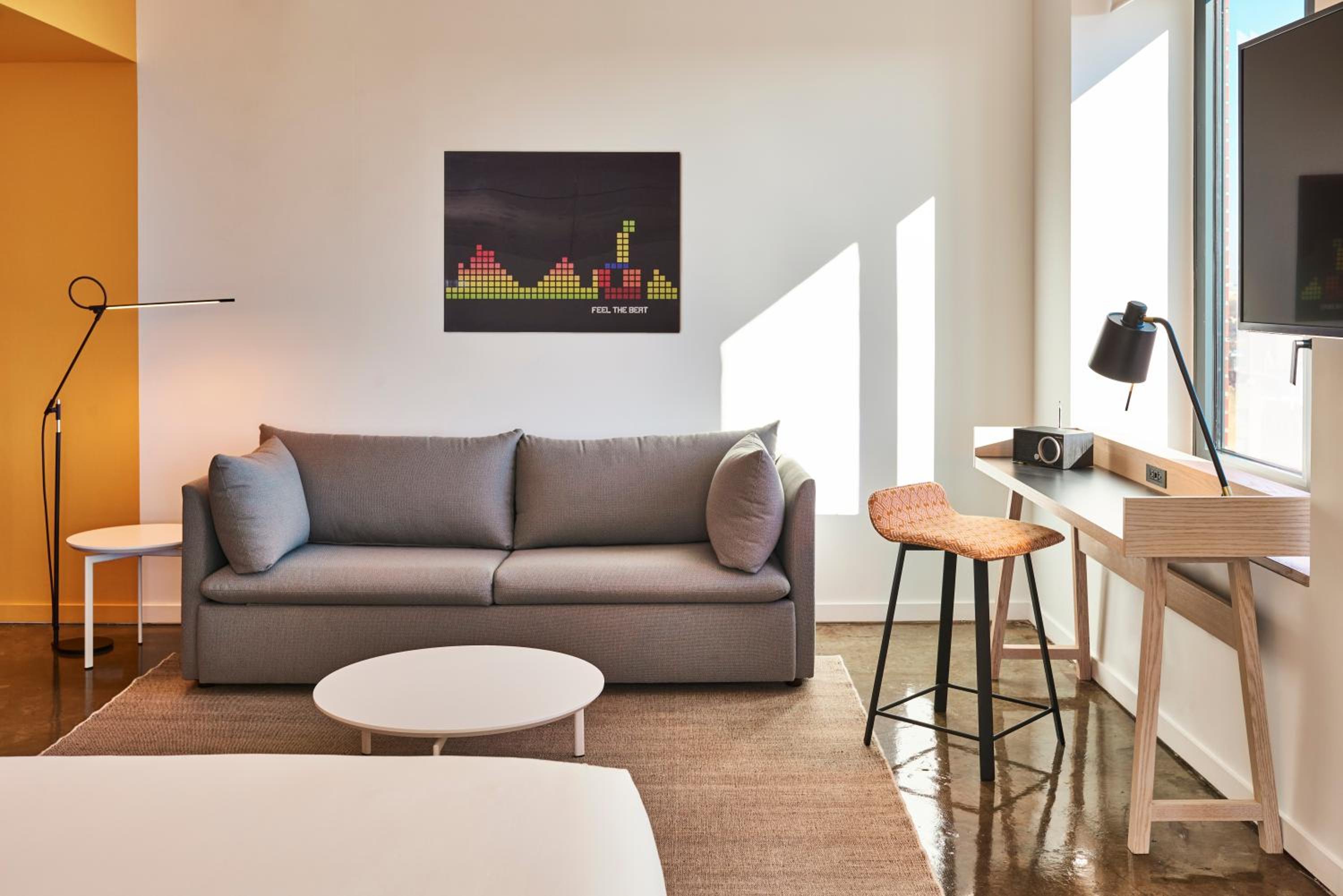 Hotel room seating area with a gray couch, small coffee table, writing desk, and colorful wall art.