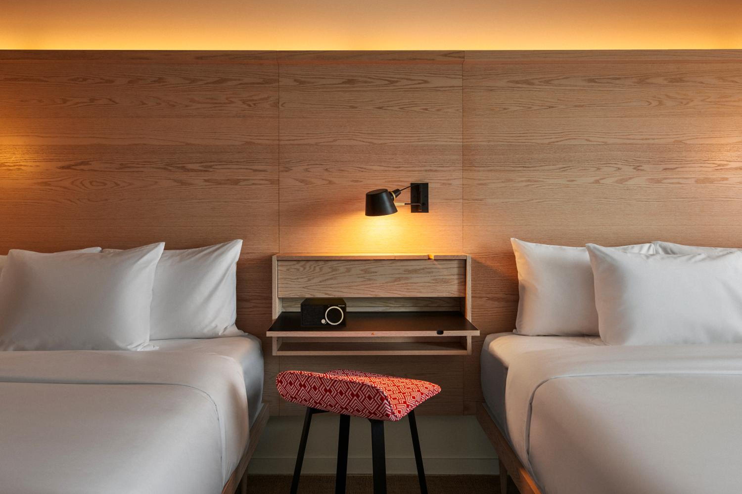 Hotel bedroom featuring beds with white linens flanking a wood panel wall with a sconce and floating shelf.