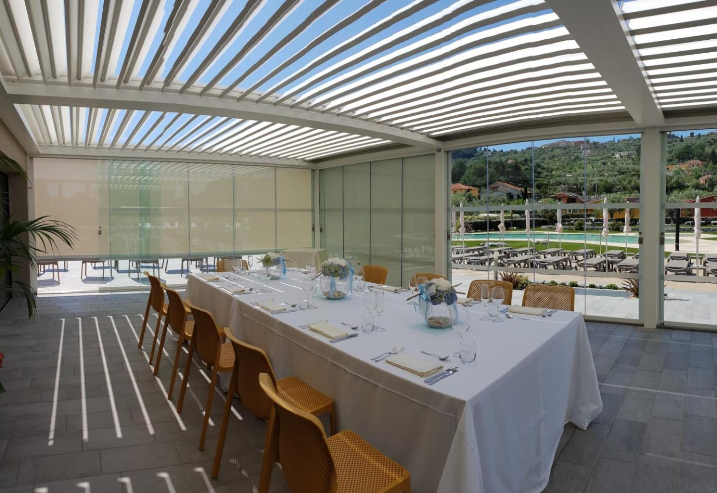 A long banquet table set with white linen under a louvered pergola enclosing a terrace with glass walls.
