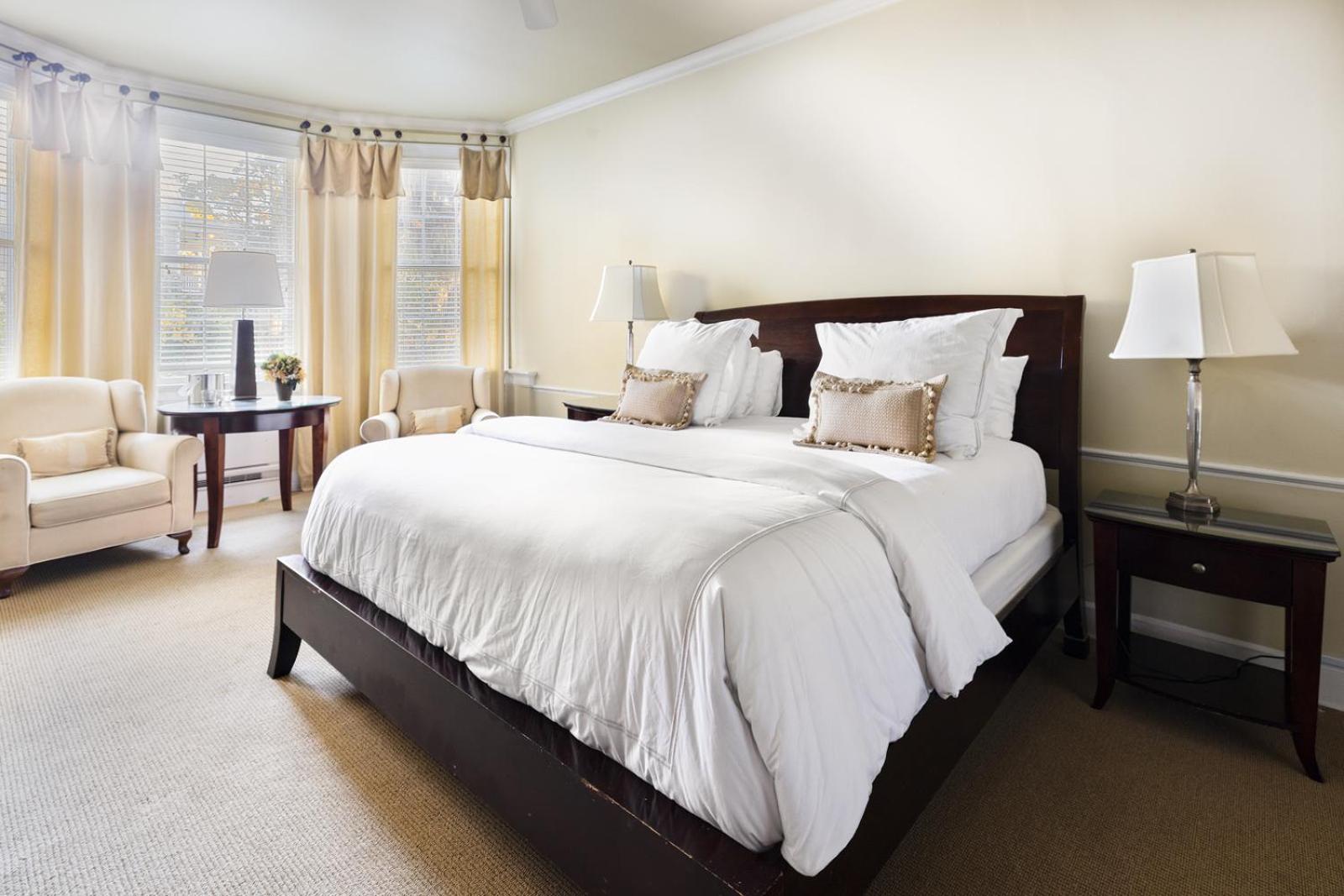 A bedroom with a dark wood bed frame, white bedding, and armchairs beside curtained bay windows.