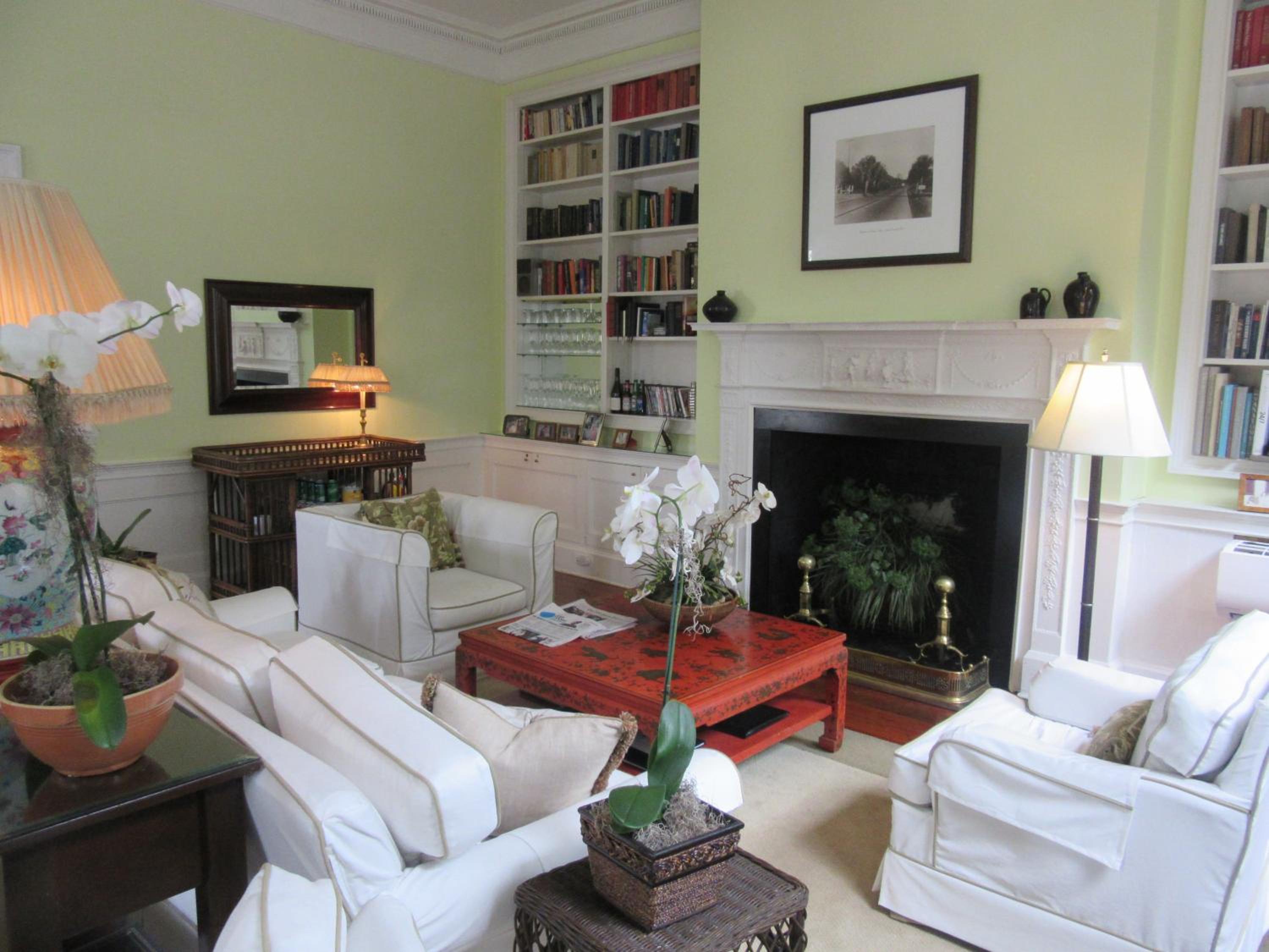 A parlor with pale green walls, a fireplace mantel, built-in bookshelves, and white slipcovered armchairs.