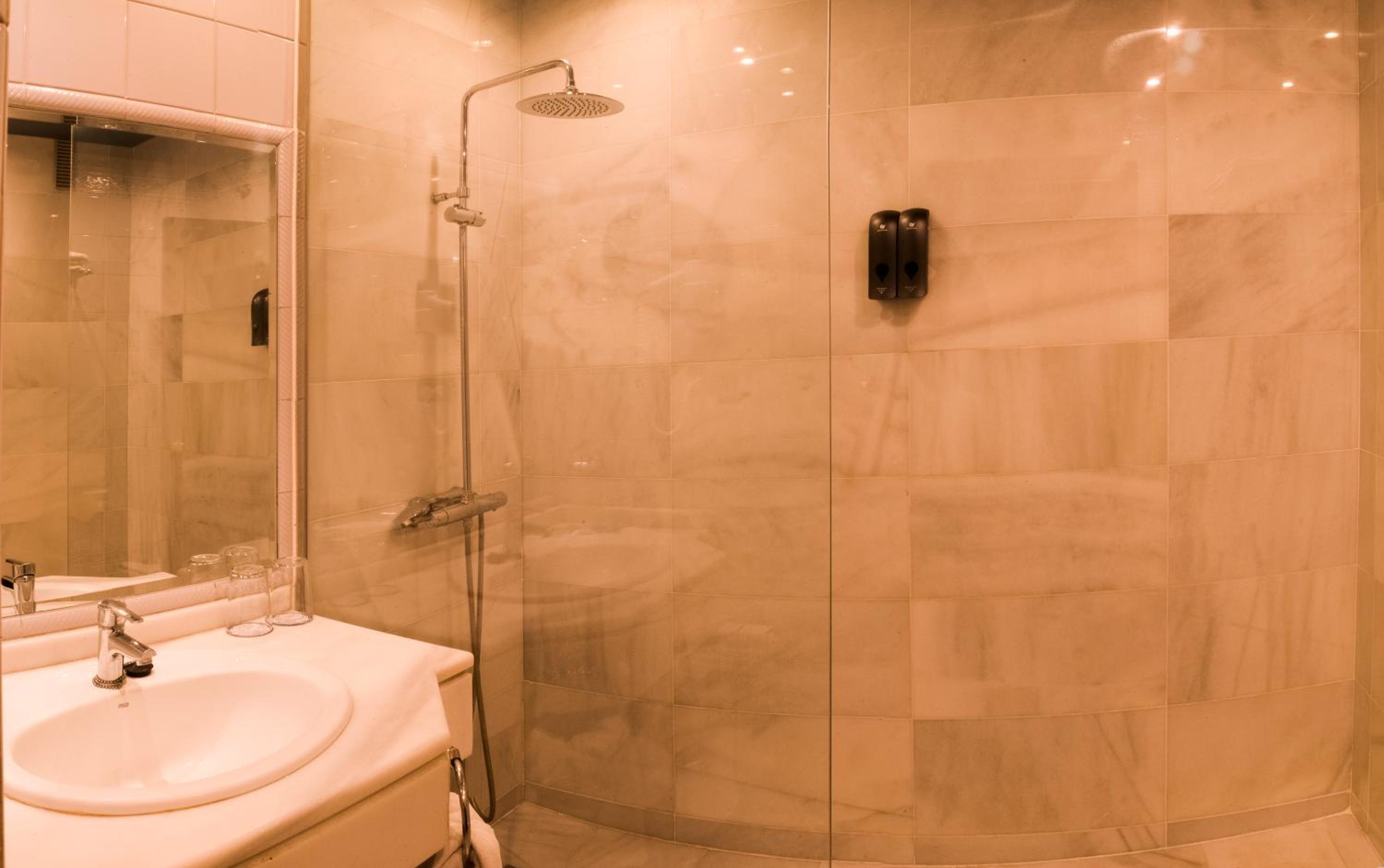 A marble-tiled bathroom with a glass walk-in shower, wall-mounted dispensers, and a white vanity sink.