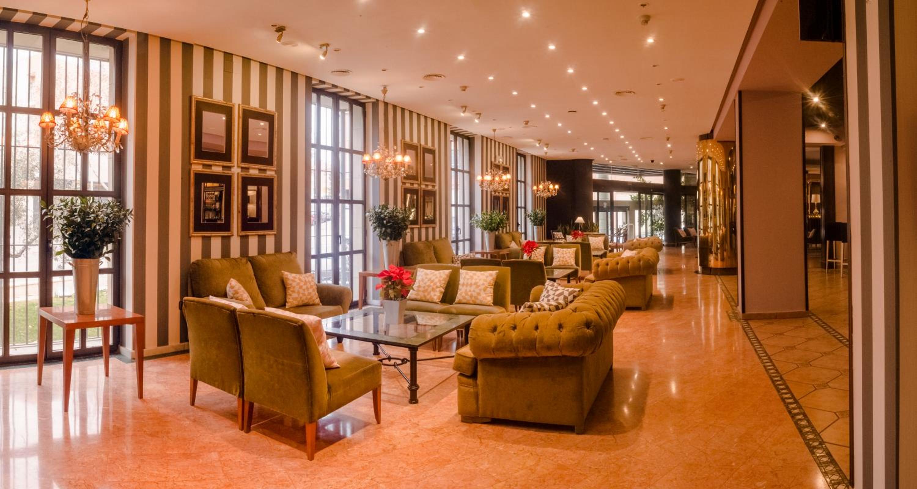 A hotel lounge with striped walls, green upholstered armchairs, glass coffee tables, chandeliers, and marble floors.