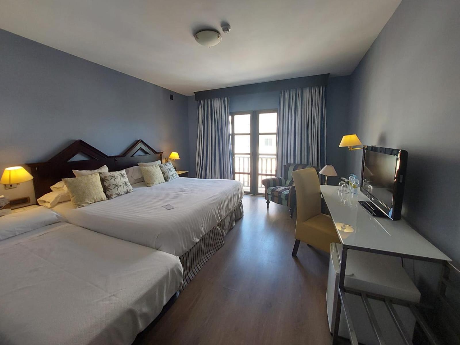 A blue-walled hotel room with wooden headboards, made beds, a desk with television, and curtained balcony doors.
