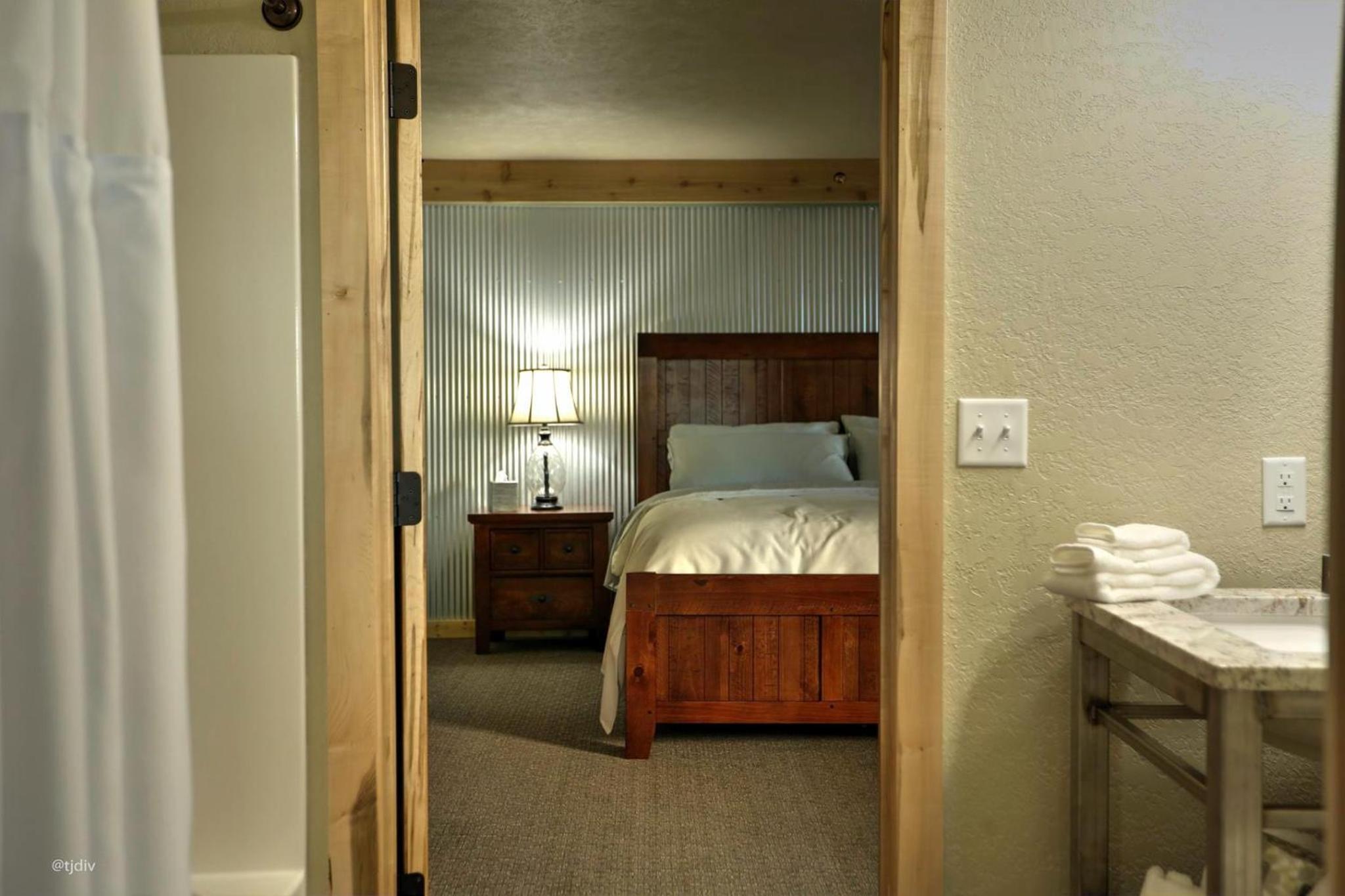 View through a bathroom doorway to a wooden bed with white bedding against a corrugated metal wall.