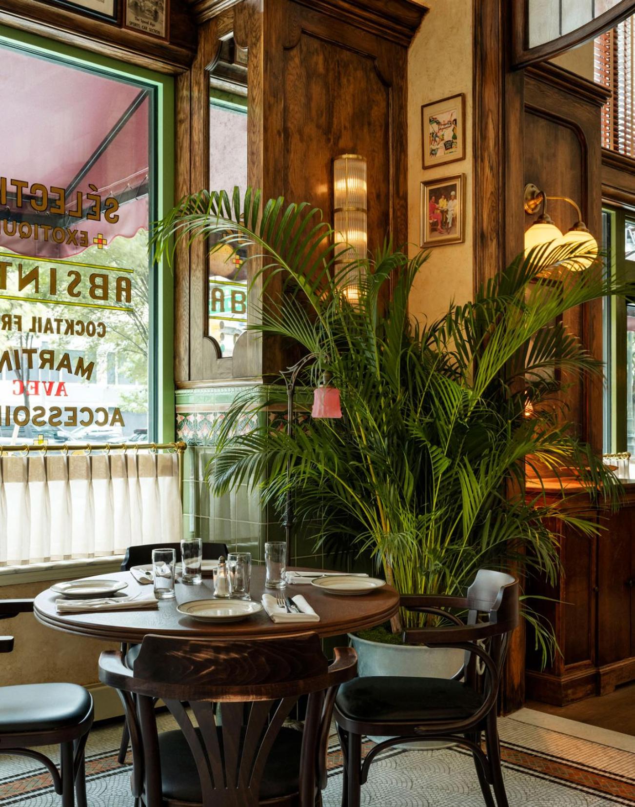 A bistro corner with rich wood paneling, a leafy potted palm, and a round wooden dining table by a window.
