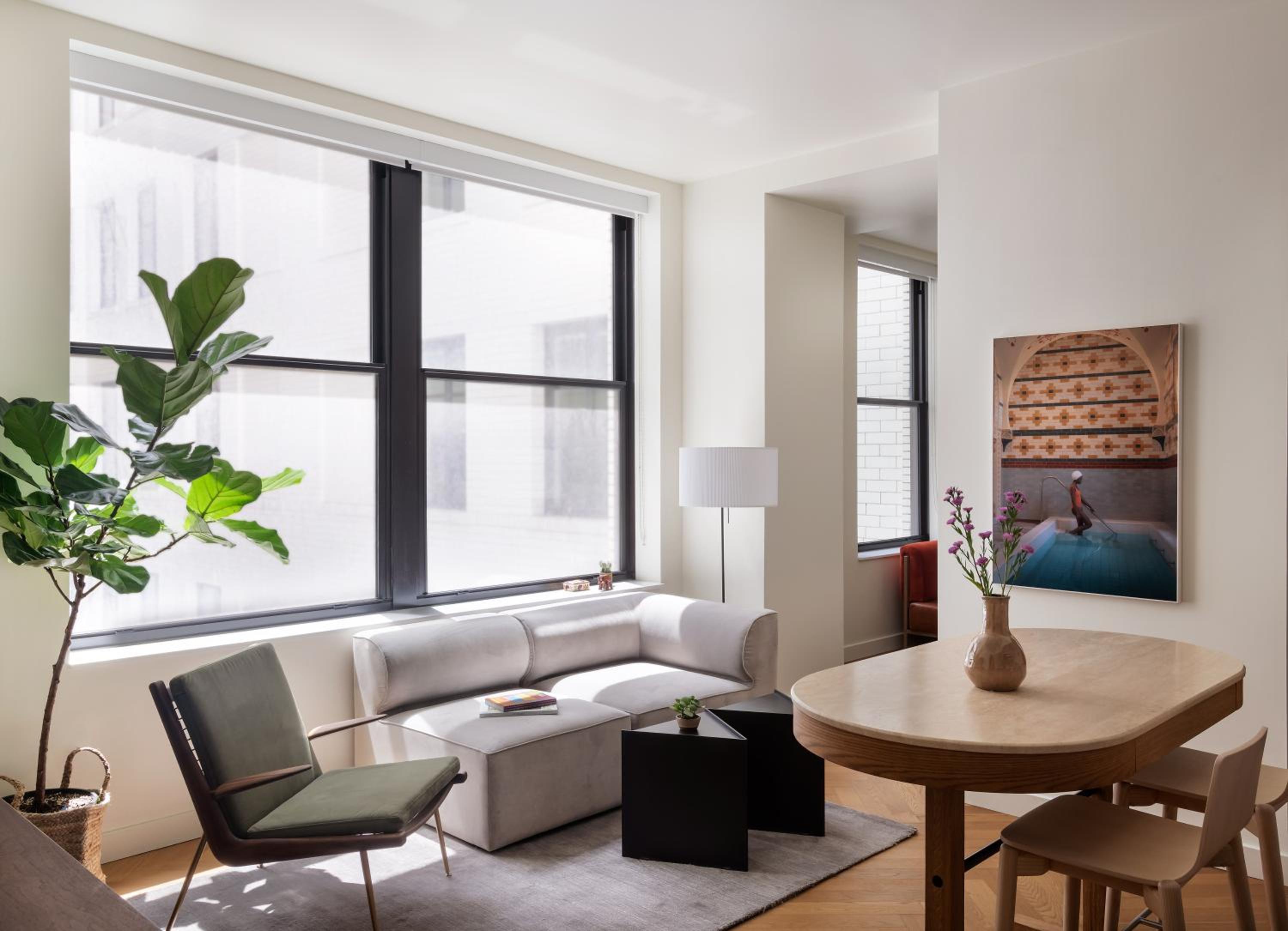 A sunlit apartment interior with large black-framed windows, a low beige sectional, and a pale wood dining table.