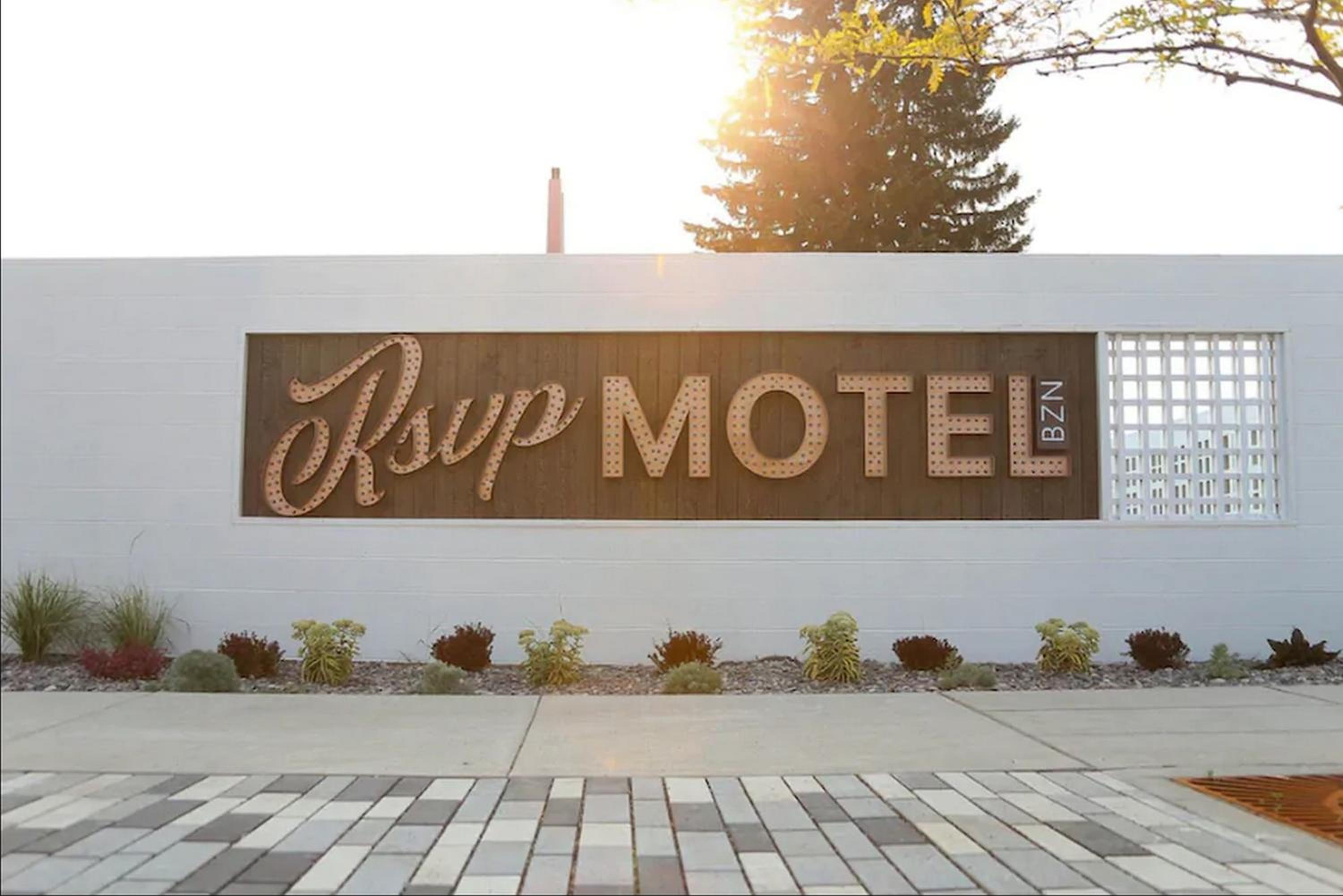 A backlit sign for RSVP Motel mounted on a white brick wall above low plantings and paved sidewalk.