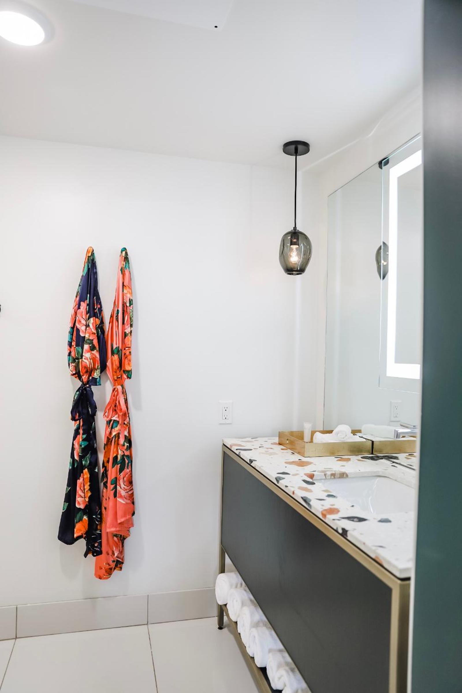 Floral robes hang on the wall beside a bathroom vanity with a speckled countertop and illuminated mirror.