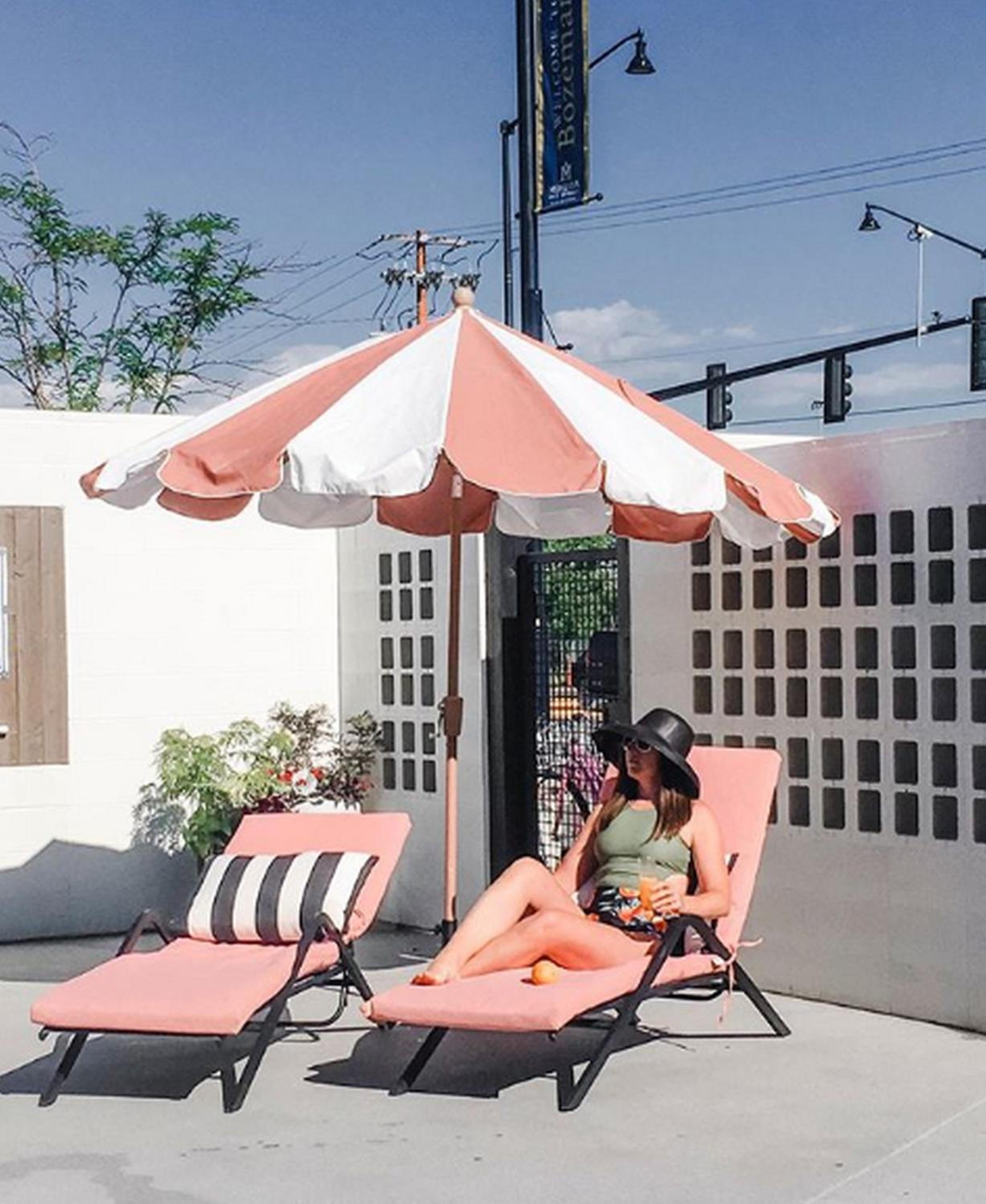 A woman relaxes on a pink sun lounger beneath a striped umbrella beside a breeze block wall.