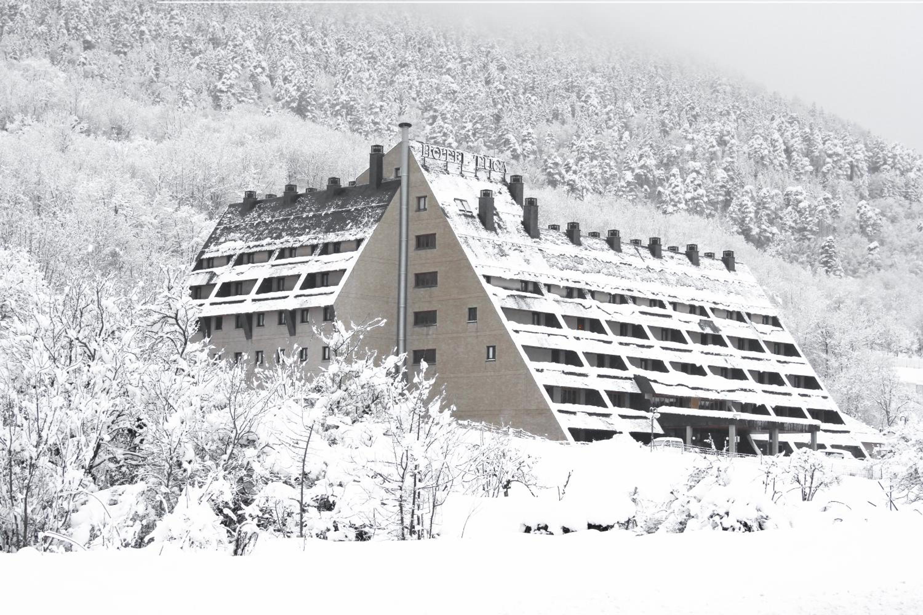 Snow blankets the triangular facade and roof of Hotel Tuca, surrounded by snow-covered pine forest.