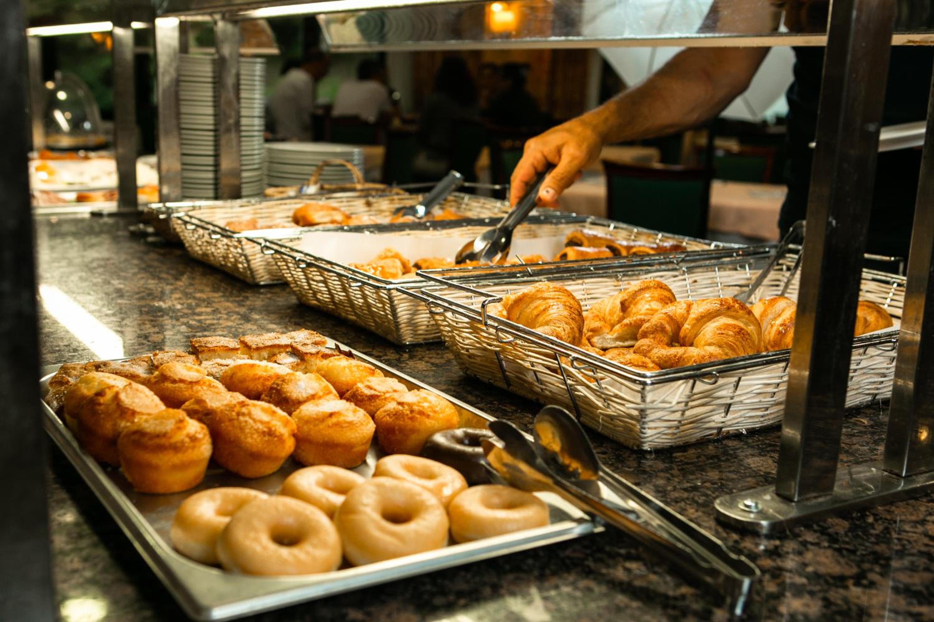 Trays of glazed donuts, pastries, and baskets of croissants arranged on a breakfast buffet counter.