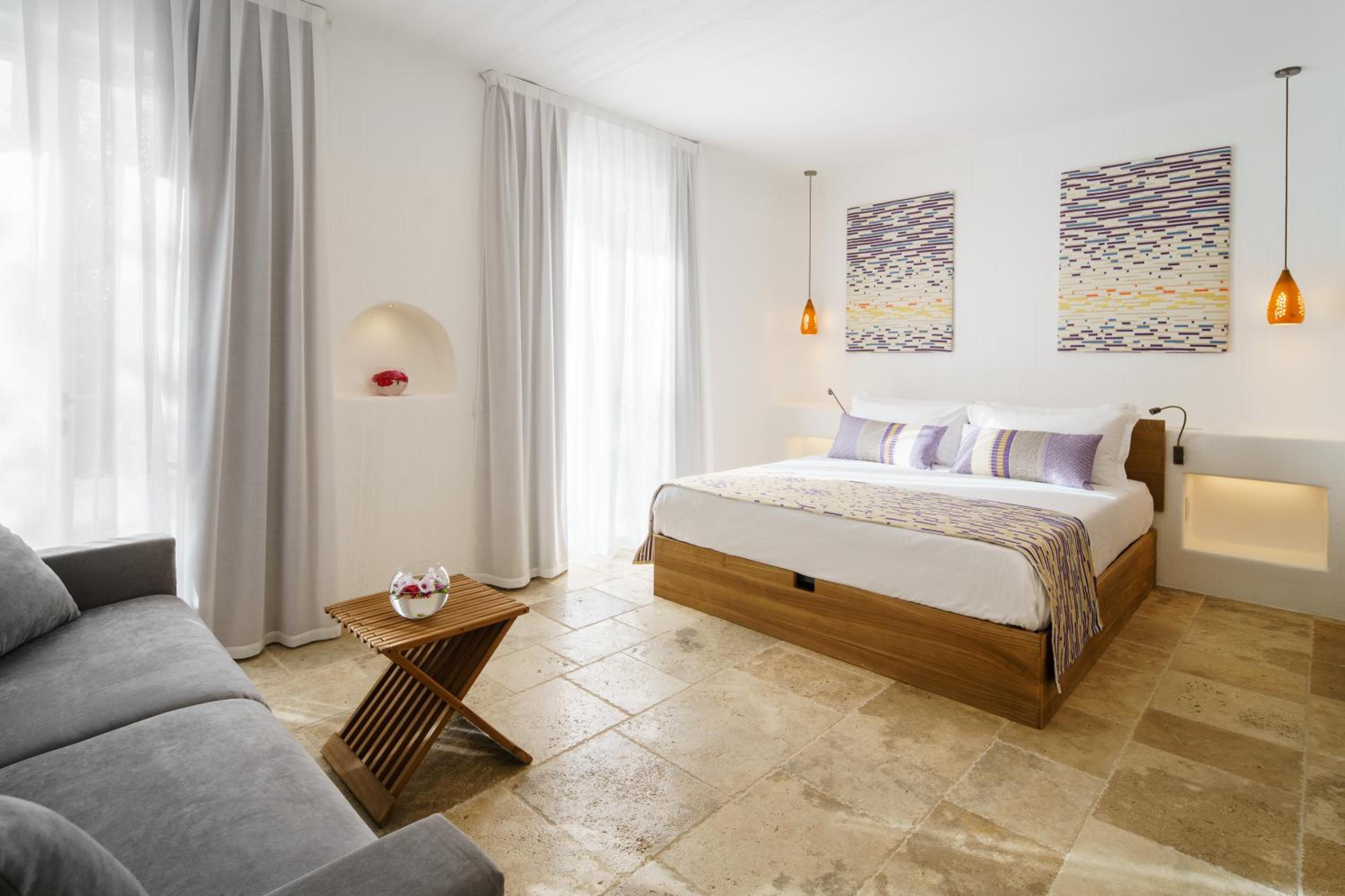 A bedroom with stone-tiled floor, a wooden platform bed with patterned pillows, and a gray sofa by sheer curtains.