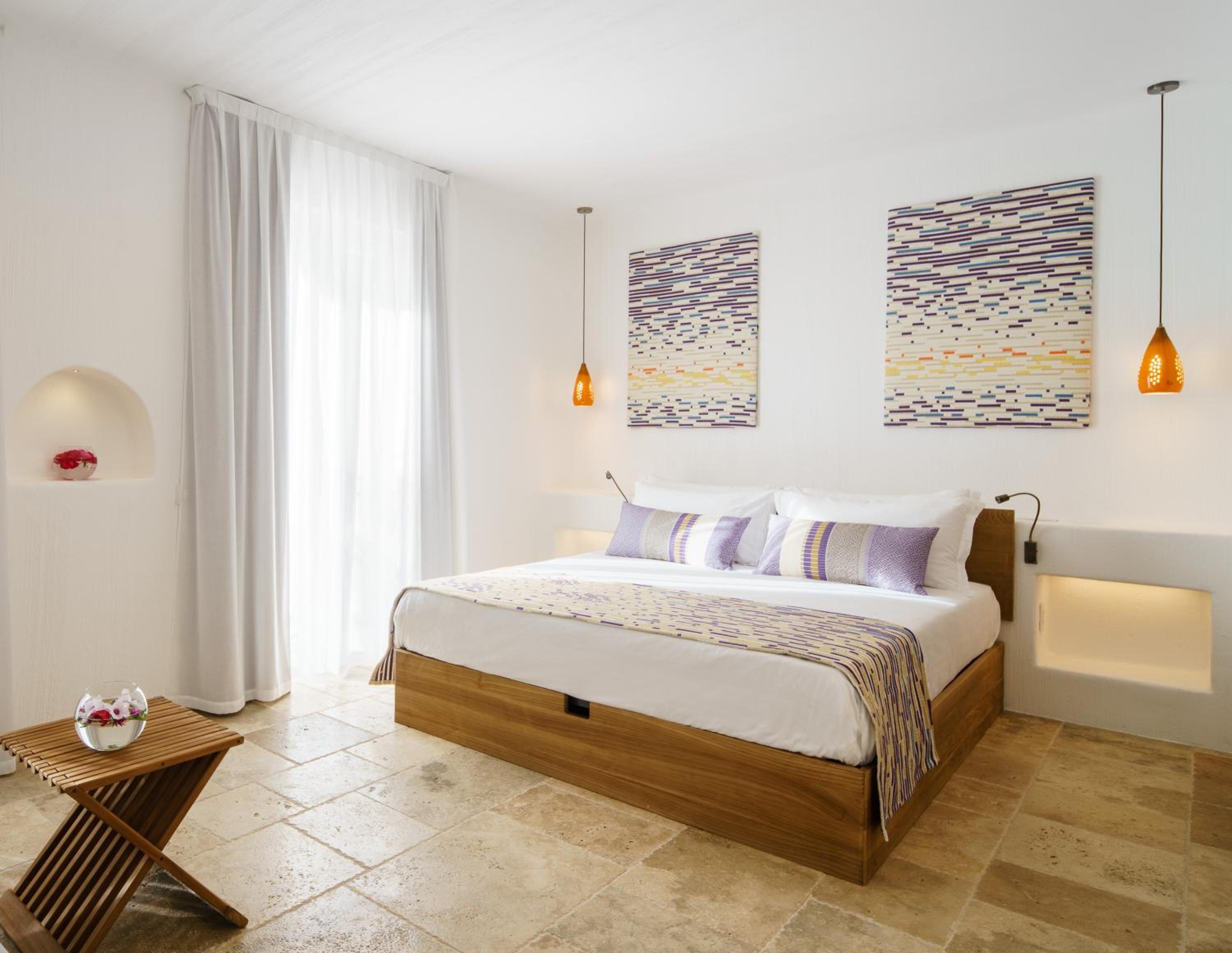 A bright bedroom with a wooden bed, textile wall art, stone tile floor, and pendant lamps casting warm light.