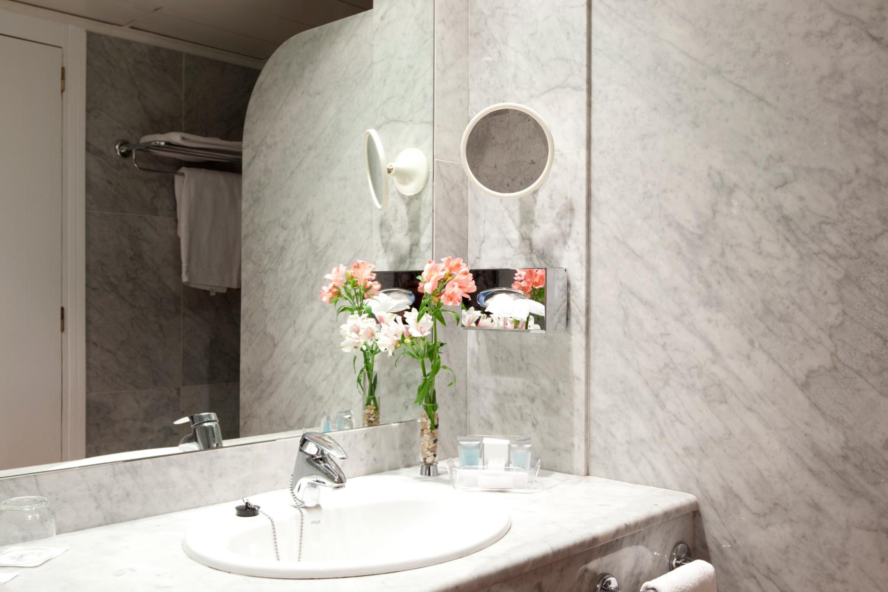 A marble bathroom vanity with an undermount sink, vase of flowers, and wall-mounted magnifying mirror.