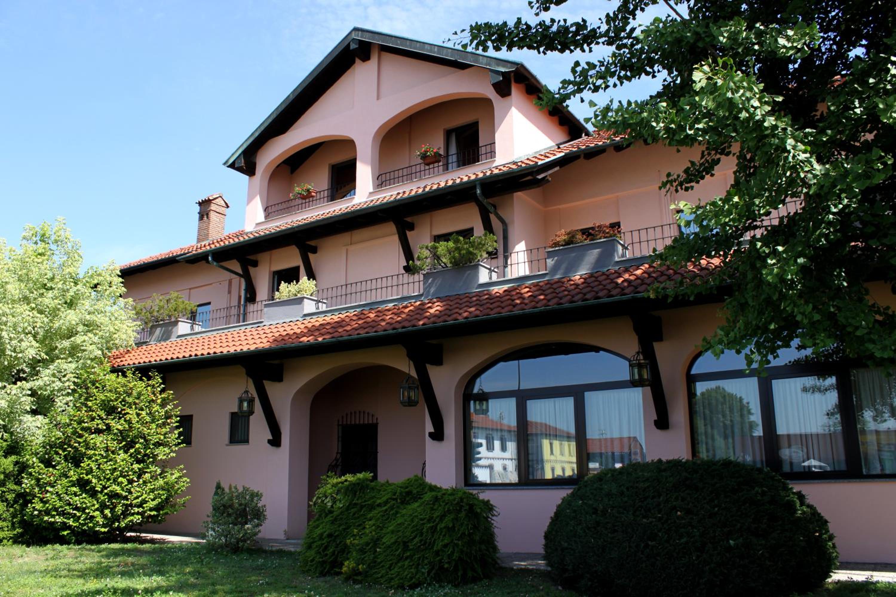 The pink facade features arched upper balconies, terracotta roof eaves, large windows, and manicured green shrubs.