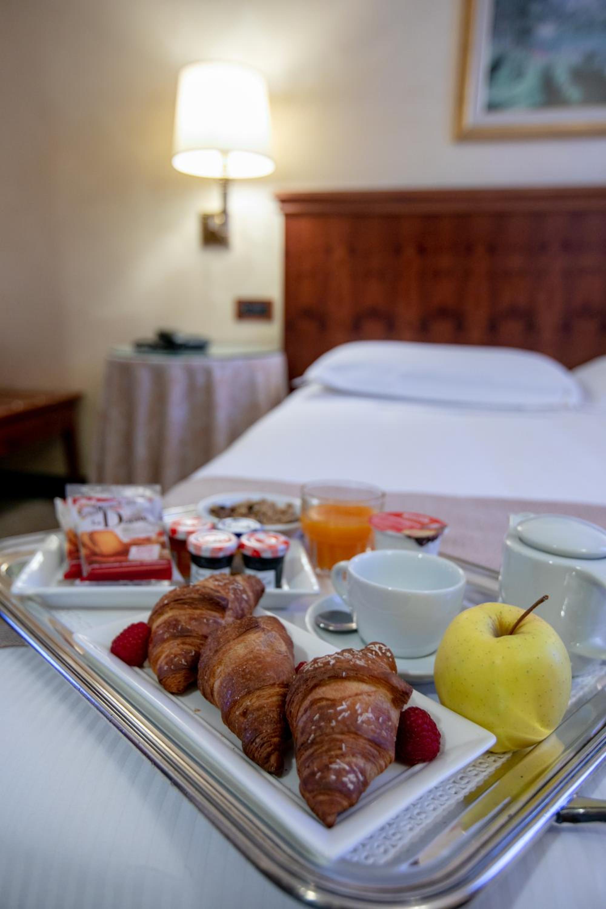 A silver breakfast tray set on a bed with croissants, fresh fruit, coffee, and packaged jams.