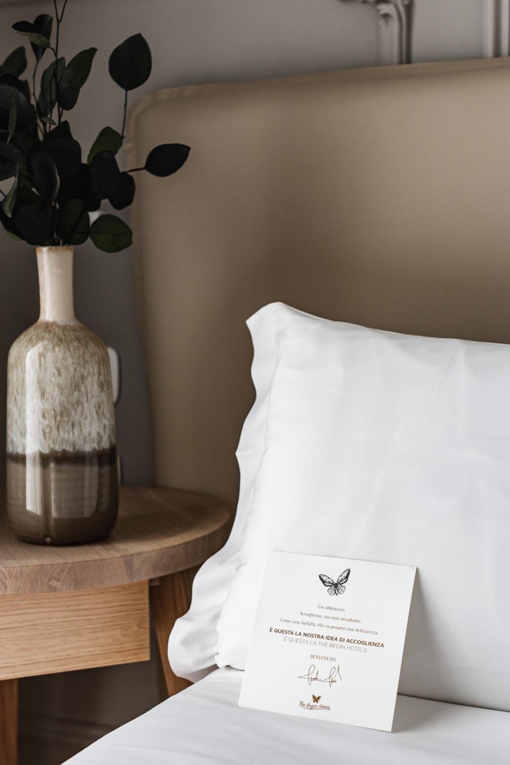 A printed welcome card rests on a white pillow next to a ceramic vase of foliage on a bedside table.