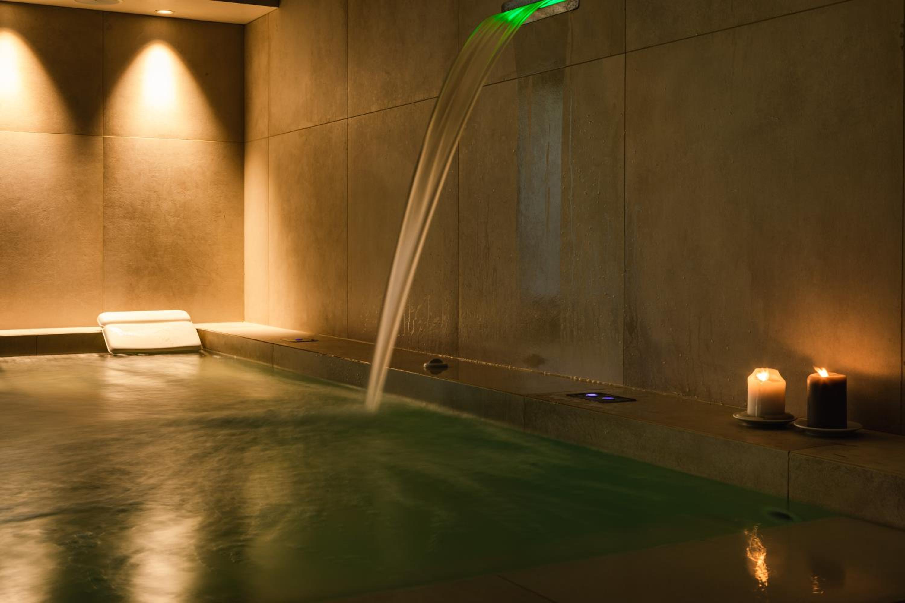 An indoor spa pool with water pouring from the wall, illuminated by recessed spotlights and lit candles.