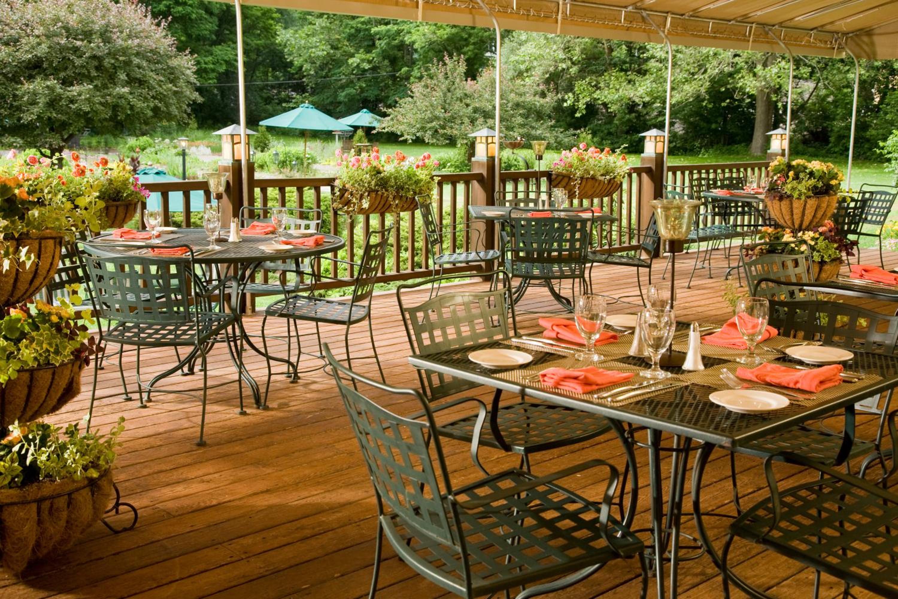 Covered wooden deck set with metal dining tables, glassware, folded napkins, and hanging flower baskets against trees.