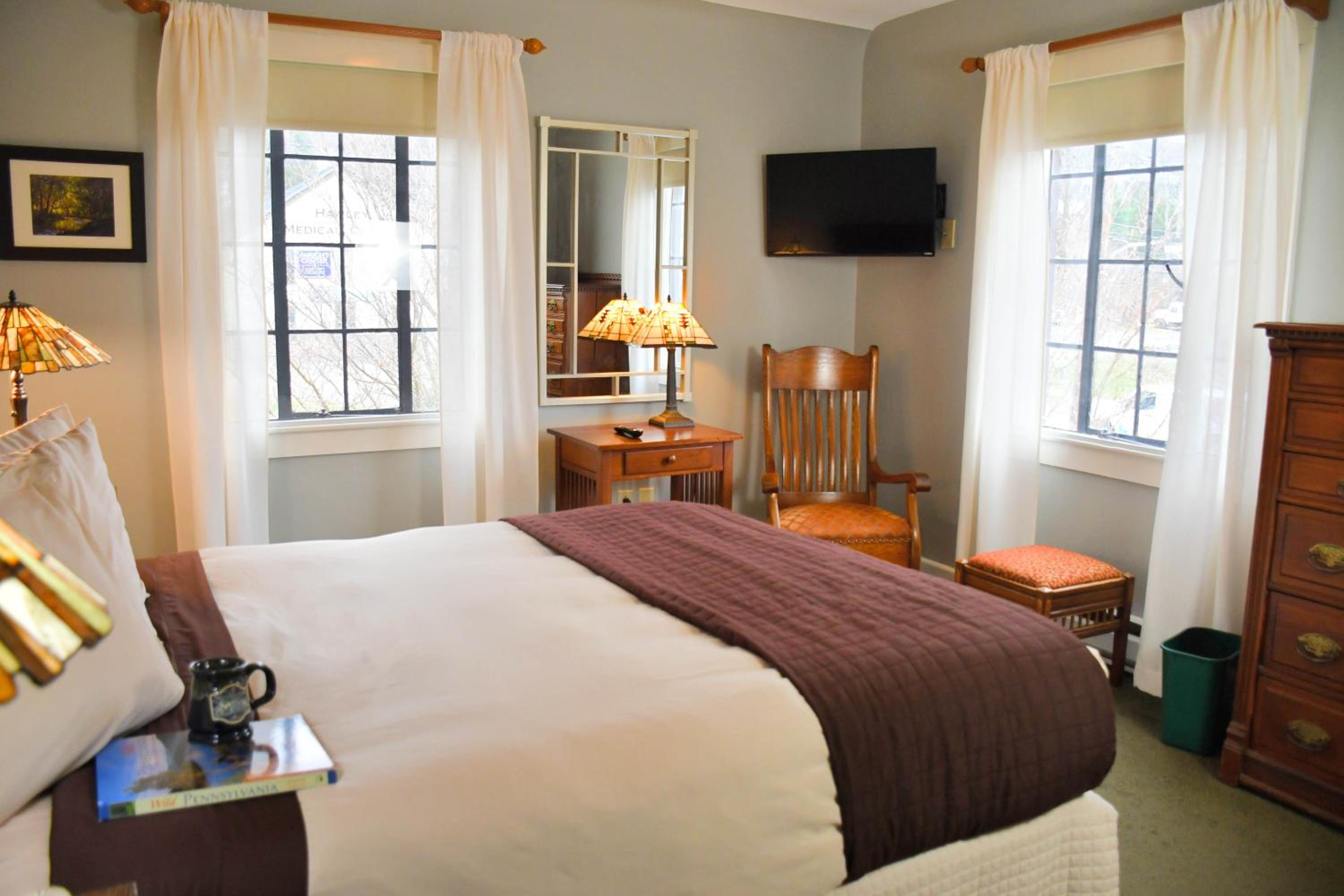Guest room with a bed, wooden rocking chair, dresser, stained glass lamps, and sheer white curtains framing windows.
