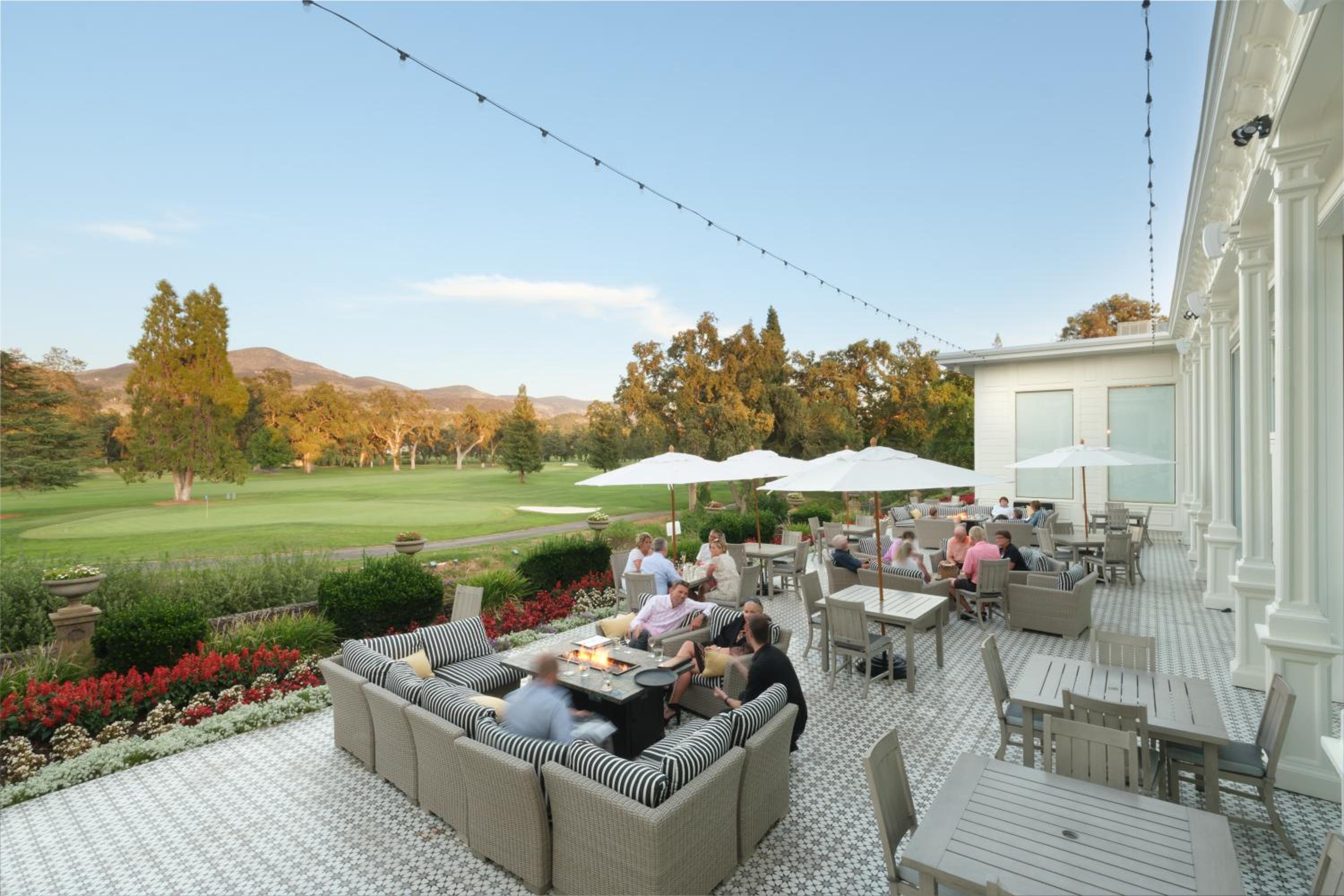 People relax on a tiled terrace with lounge chairs and umbrellas overlooking a golf course and rolling hills.
