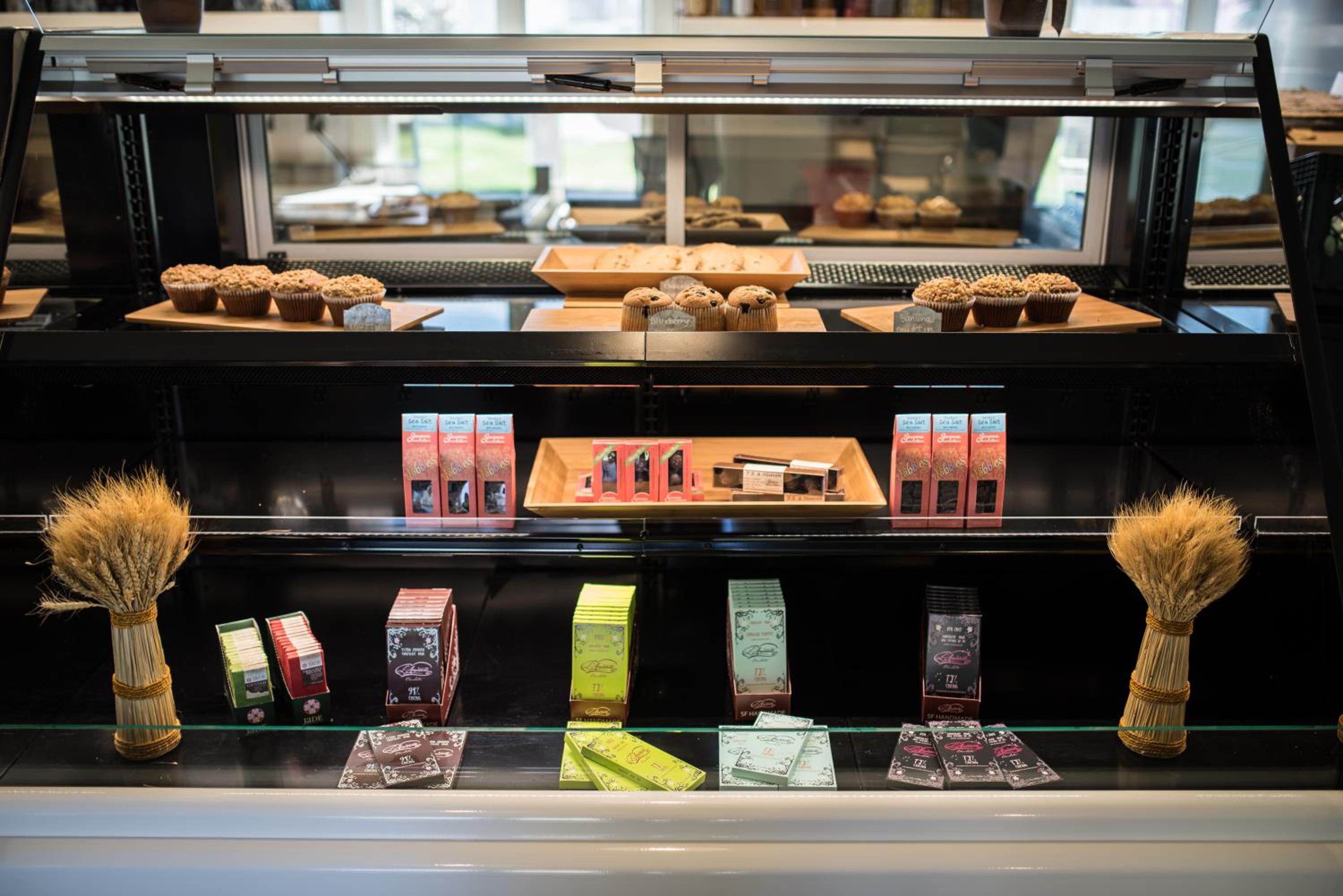 A glass display case holds fresh muffins, pastries, and artisanal chocolate bars framed by dried wheat stalks.