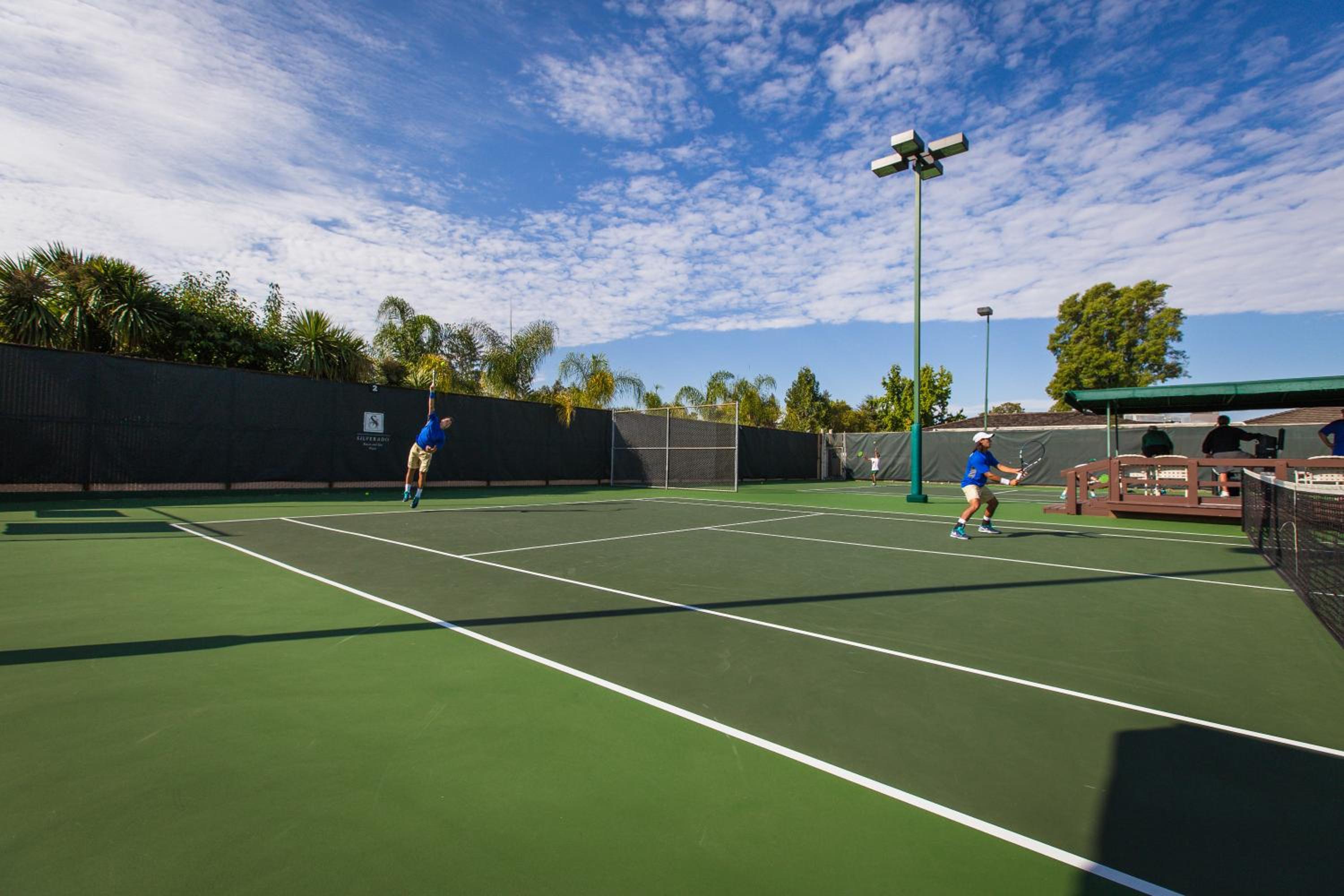 Tennis players hit balls across an outdoor green court under a partly cloudy blue sky.