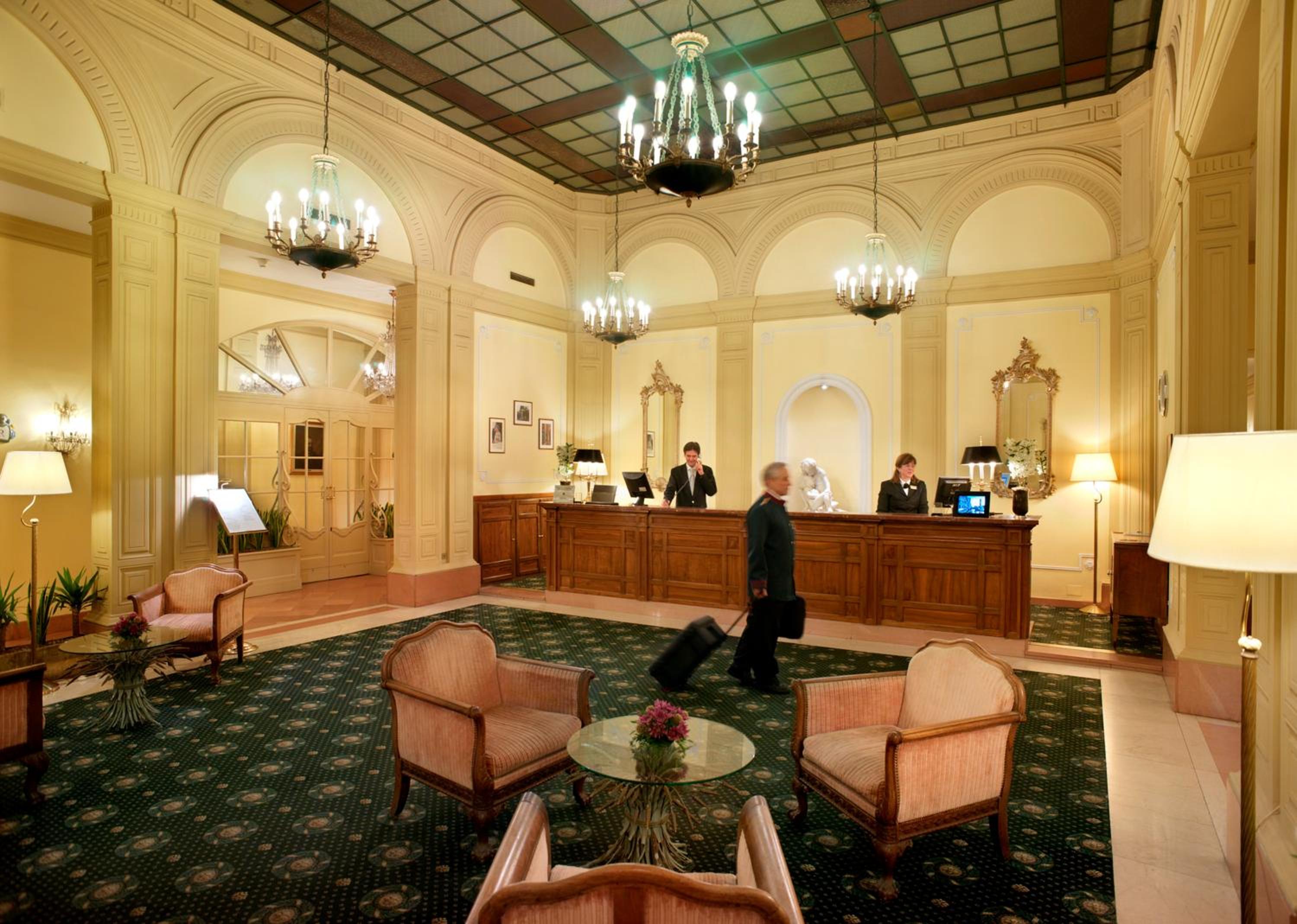 Hotel reception lobby with high arched walls, hanging chandeliers, dark patterned carpet, and a wooden counter.