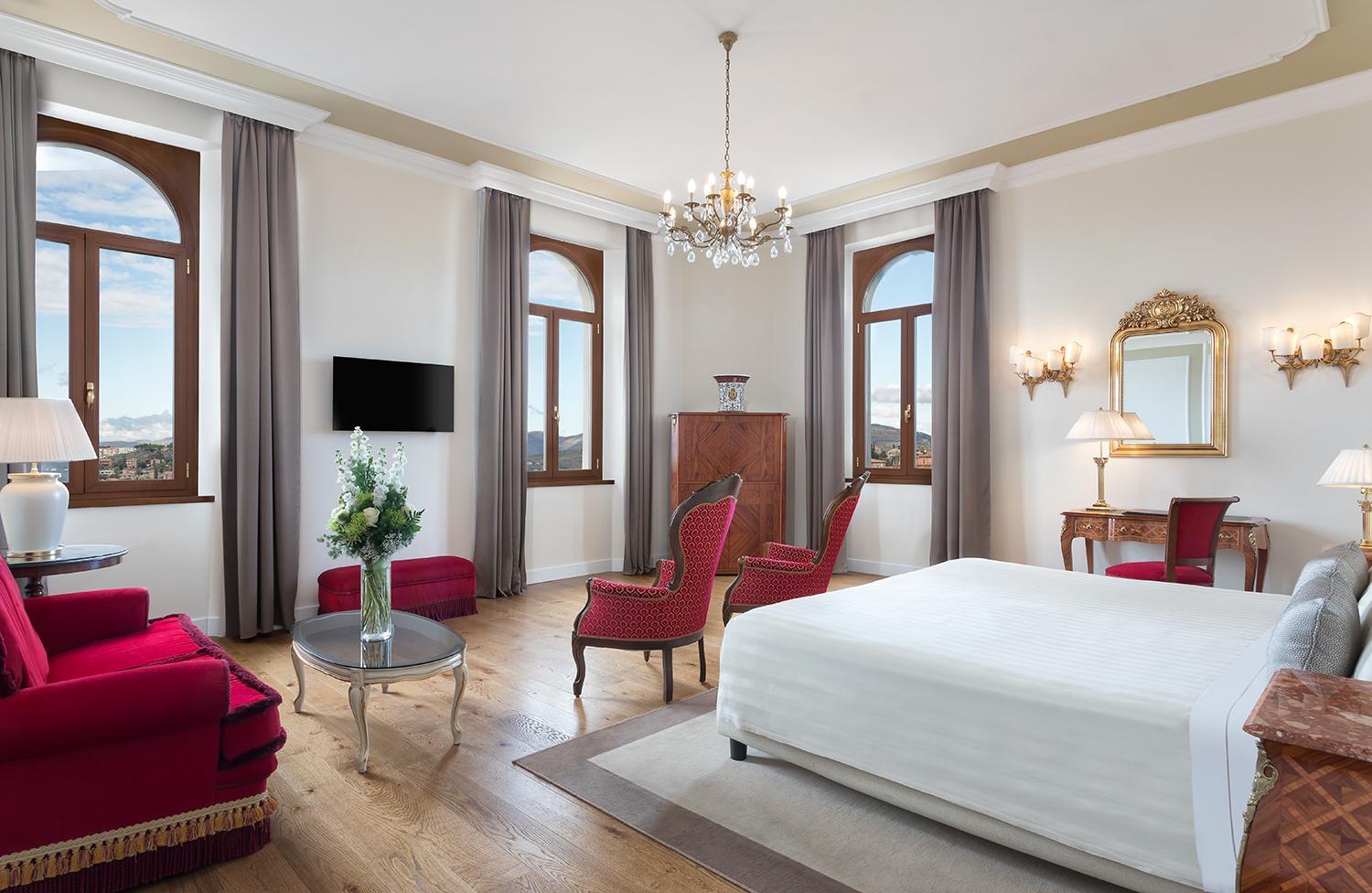 Bedroom with wood floors, tall arched windows framing the valley, a chandelier, and red armchairs.