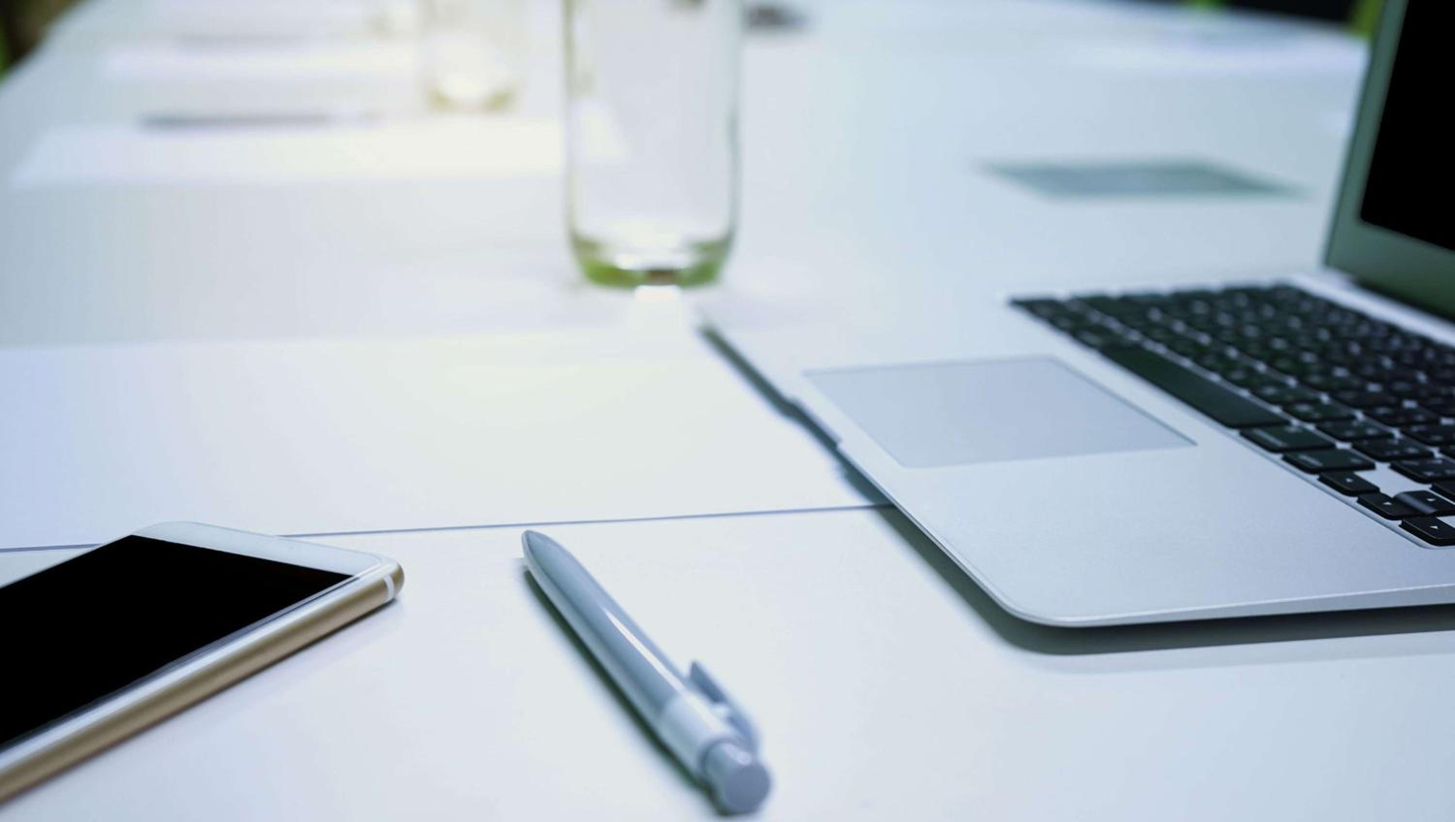 A white meeting table set with an open laptop, a pen, a smartphone, and drinking glasses.