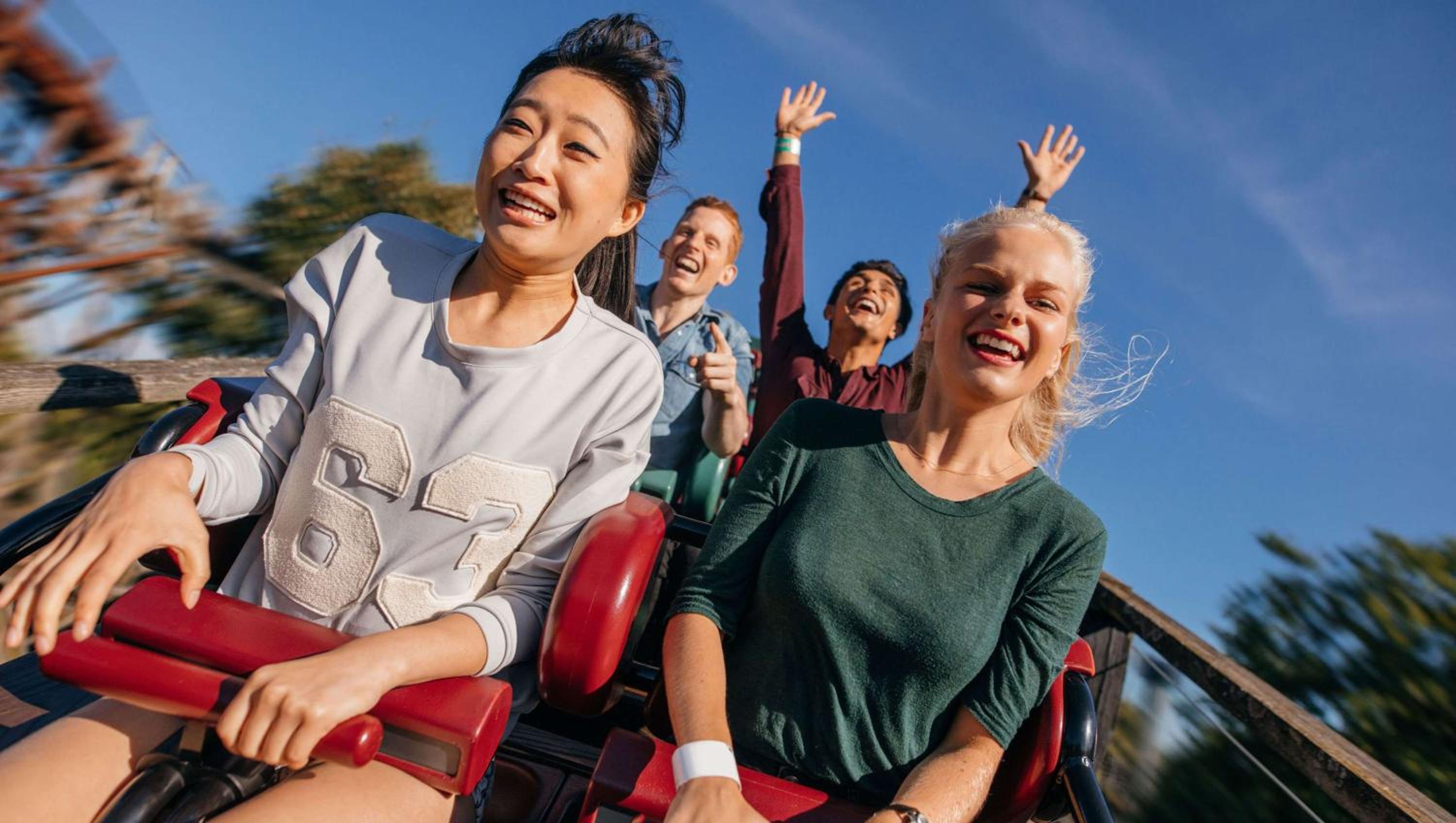 Cheerful park visitors riding a roller coaster with arms raised against a clear blue sky.