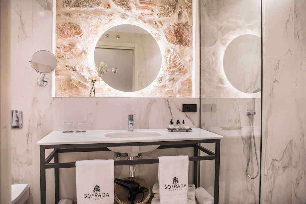 Modern bathroom featuring a marble vanity, exposed stone wall, and a circular backlit mirror next to a shower.