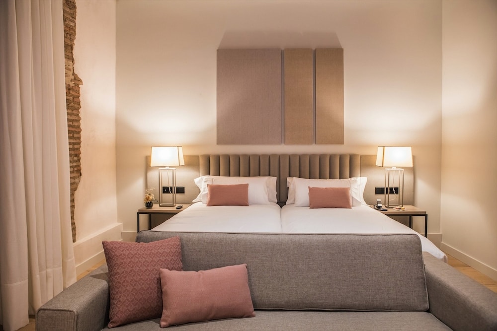 Bedroom with a gray sofa in the foreground, upholstered headboard, glowing bedside lamps, and an exposed stone wall.