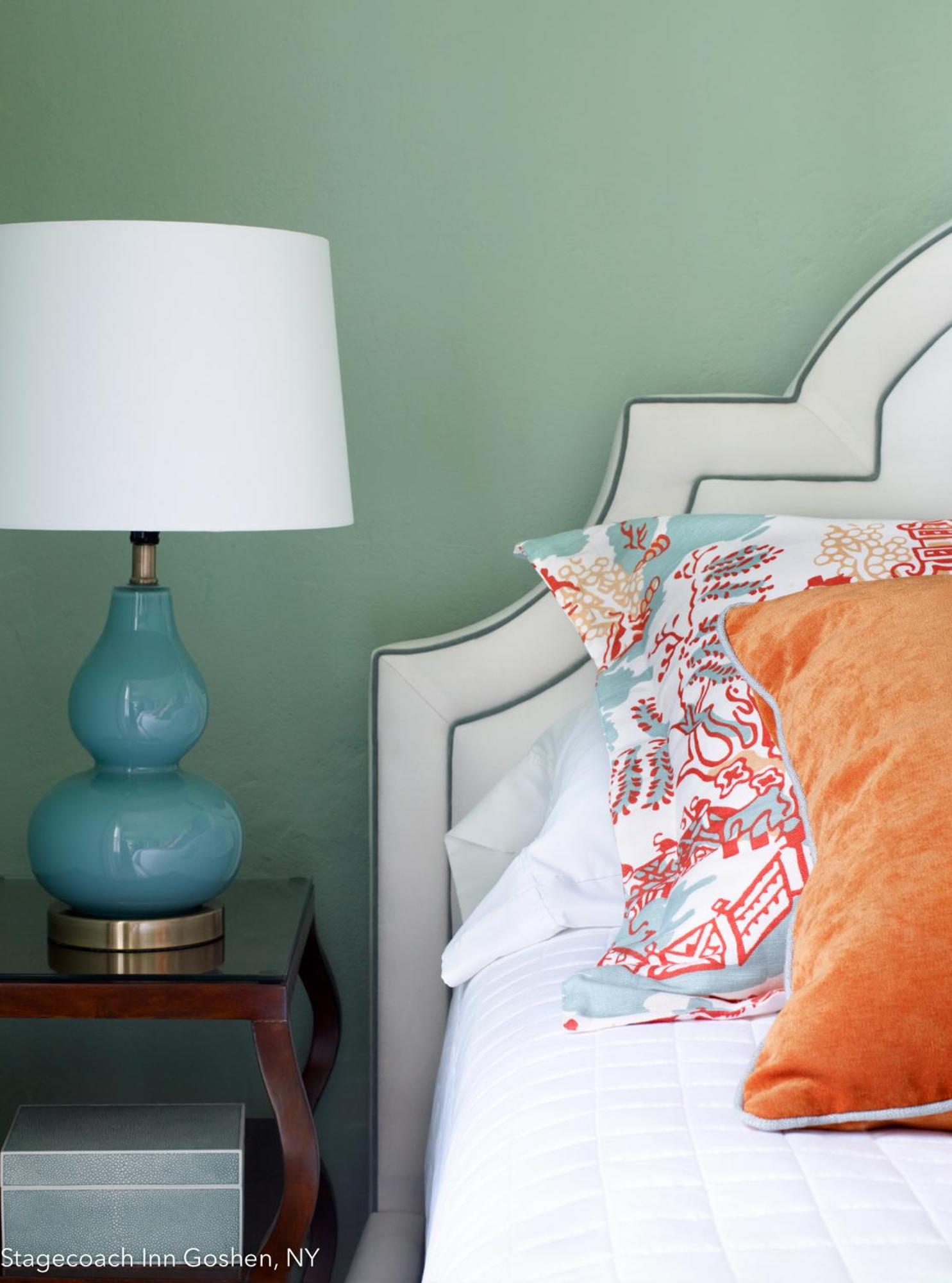 A bedroom corner with a pale green wall, an upholstered headboard, an aqua lamp, and patterned pillows.