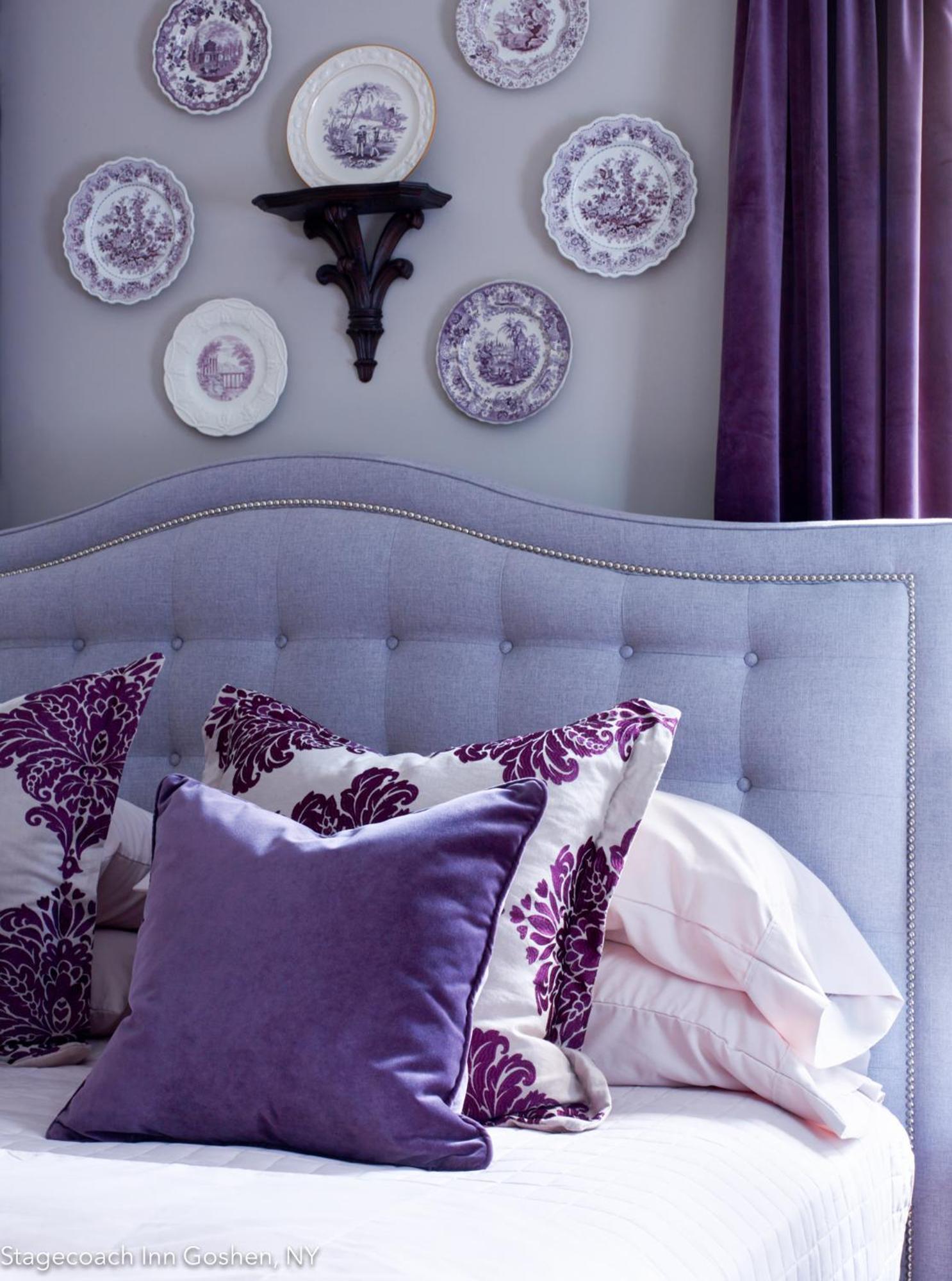 A tufted lavender headboard dressed with purple velvet pillows below decorative china plates mounted on the wall.
