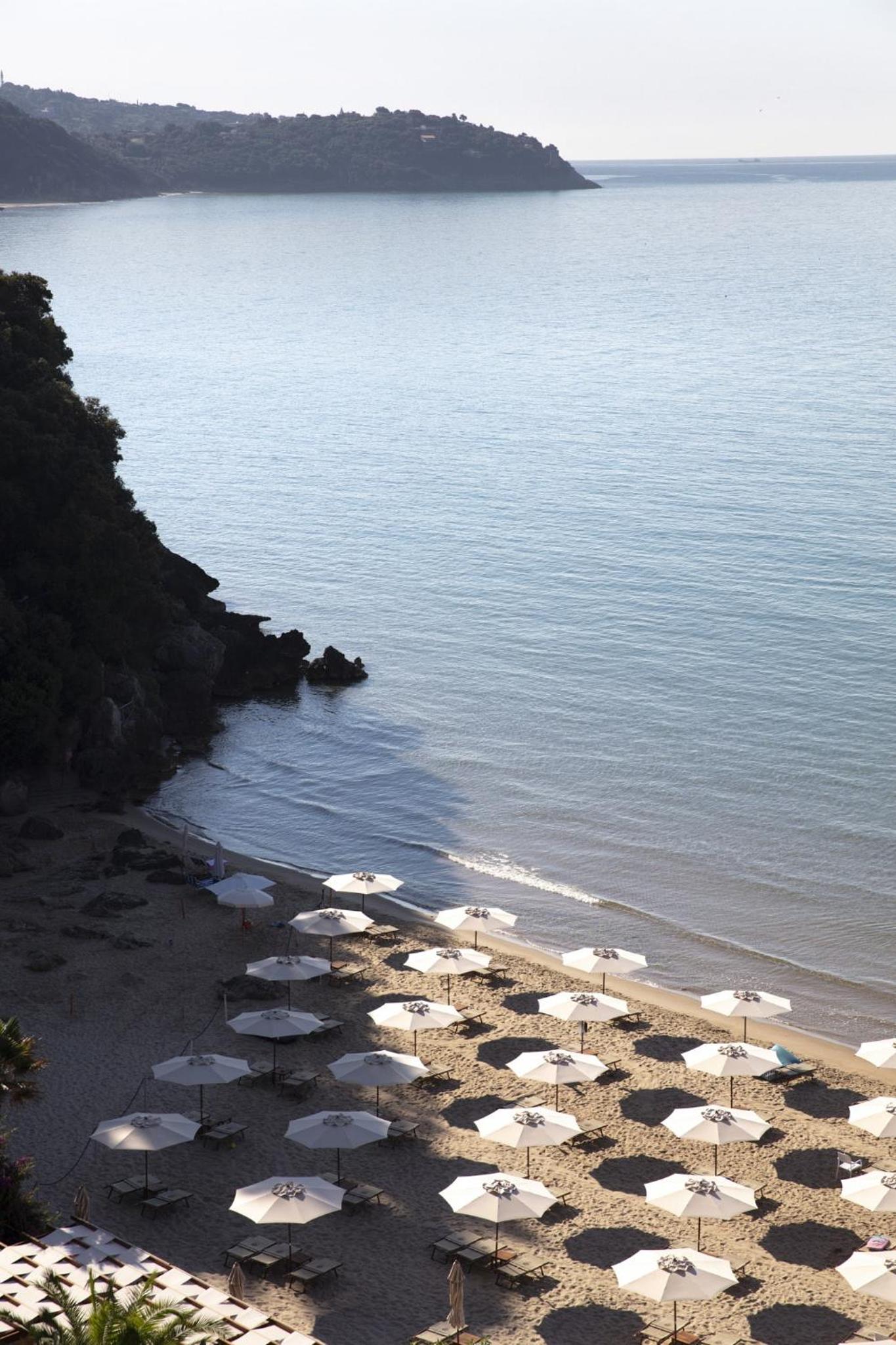 White umbrellas and lounge chairs line a sandy beach beside clear coastal waters and a rocky headland.