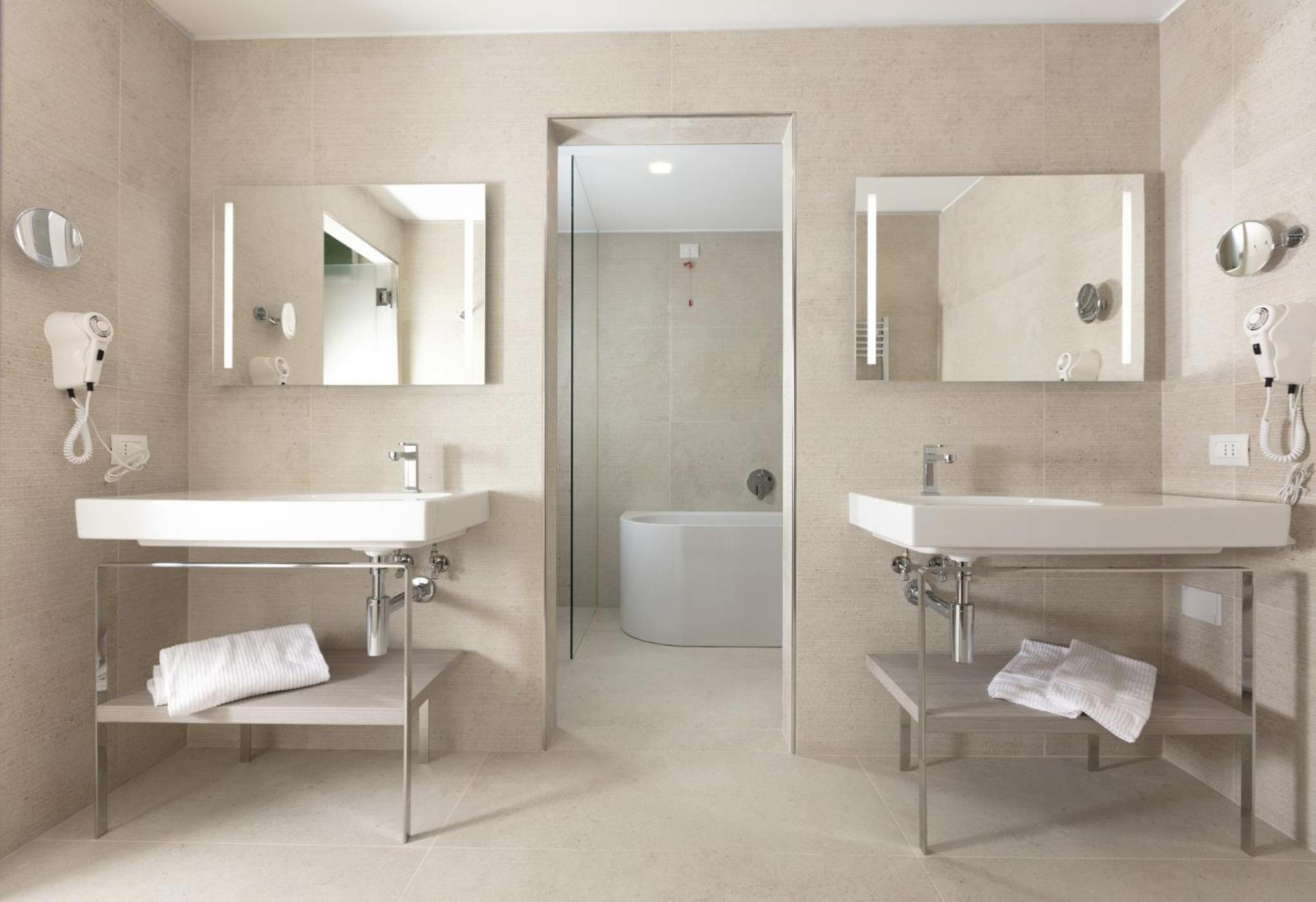 Beige tile covers a bathroom with washbasins, lighted mirrors, and a bathtub in an adjoining alcove.