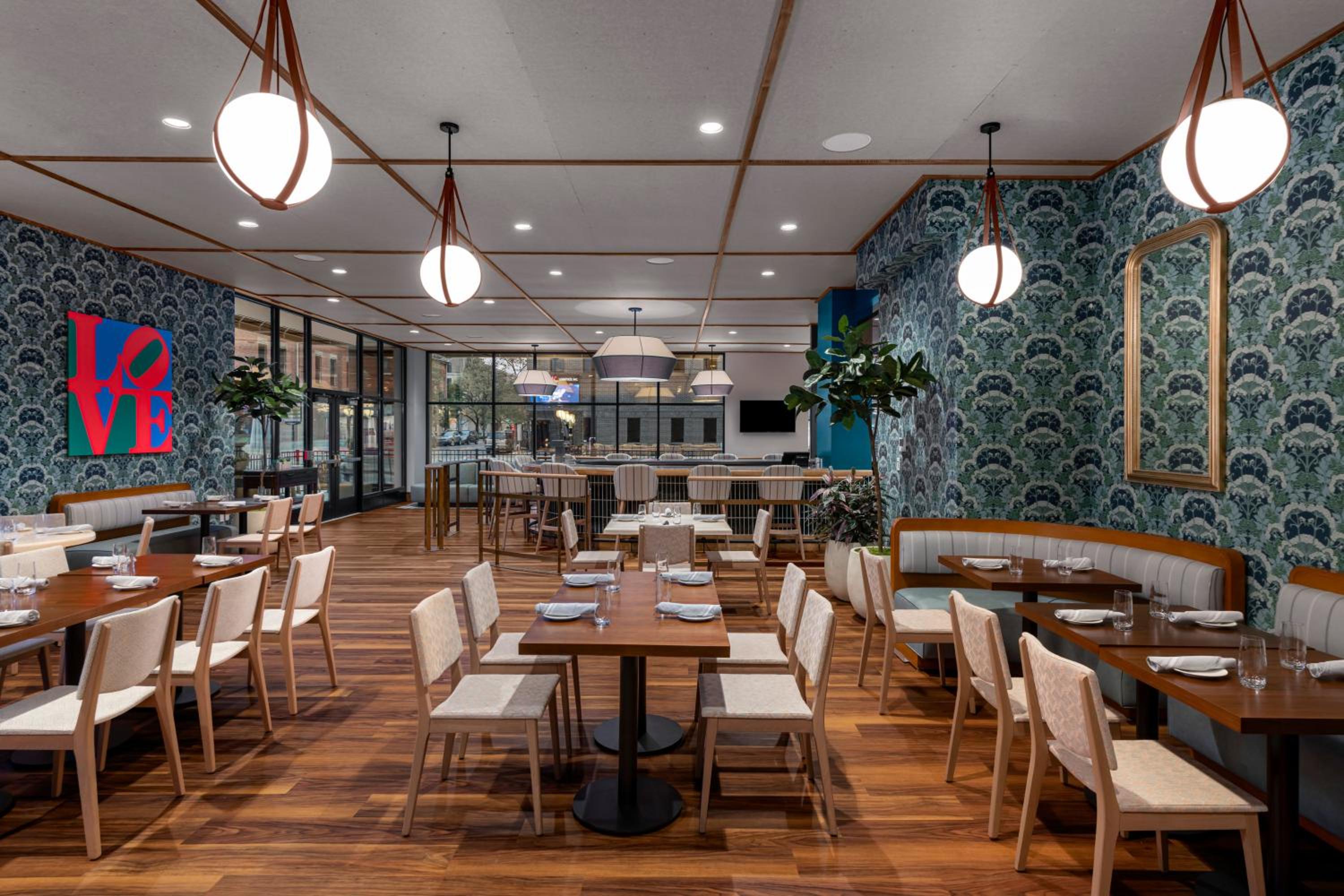 Restaurant dining room with botanical wallpaper, wooden tables and chairs, booth seating, and globe pendant lamps.