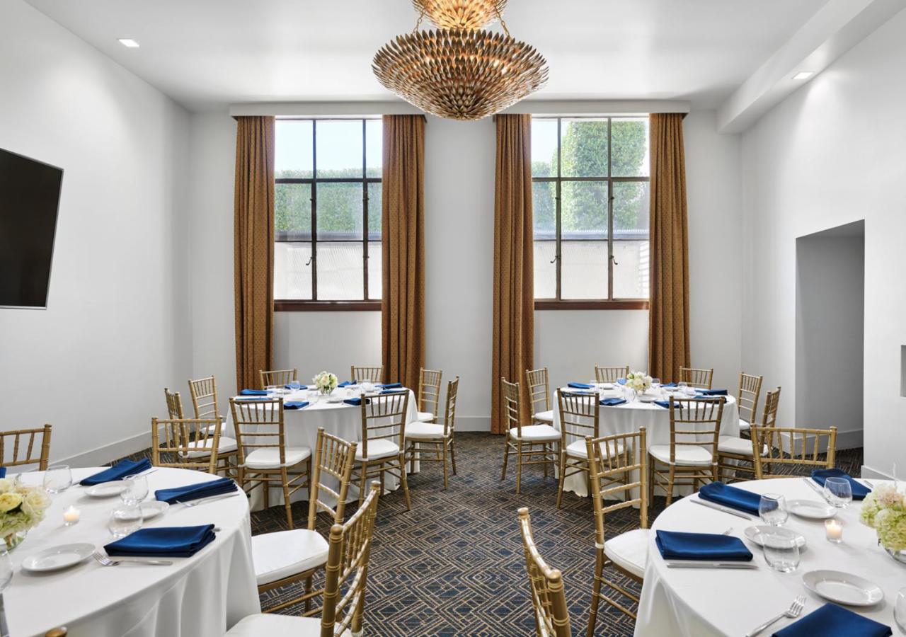 Event dining room with round tables covered in white linens, gold chairs, tall windows, and a metallic chandelier.