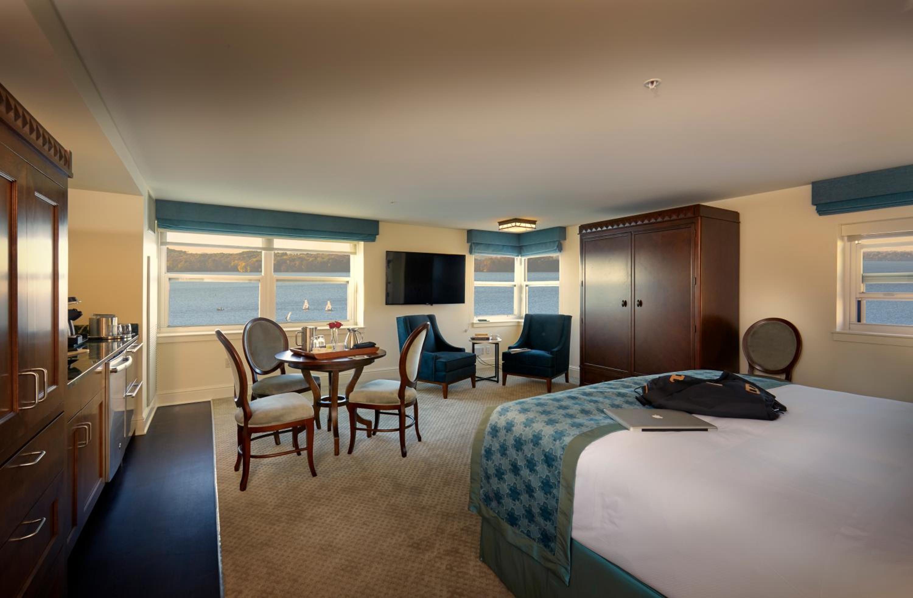 Hotel suite with a bed, small dining table, armchairs, and wide windows overlooking a blue lake in daylight.