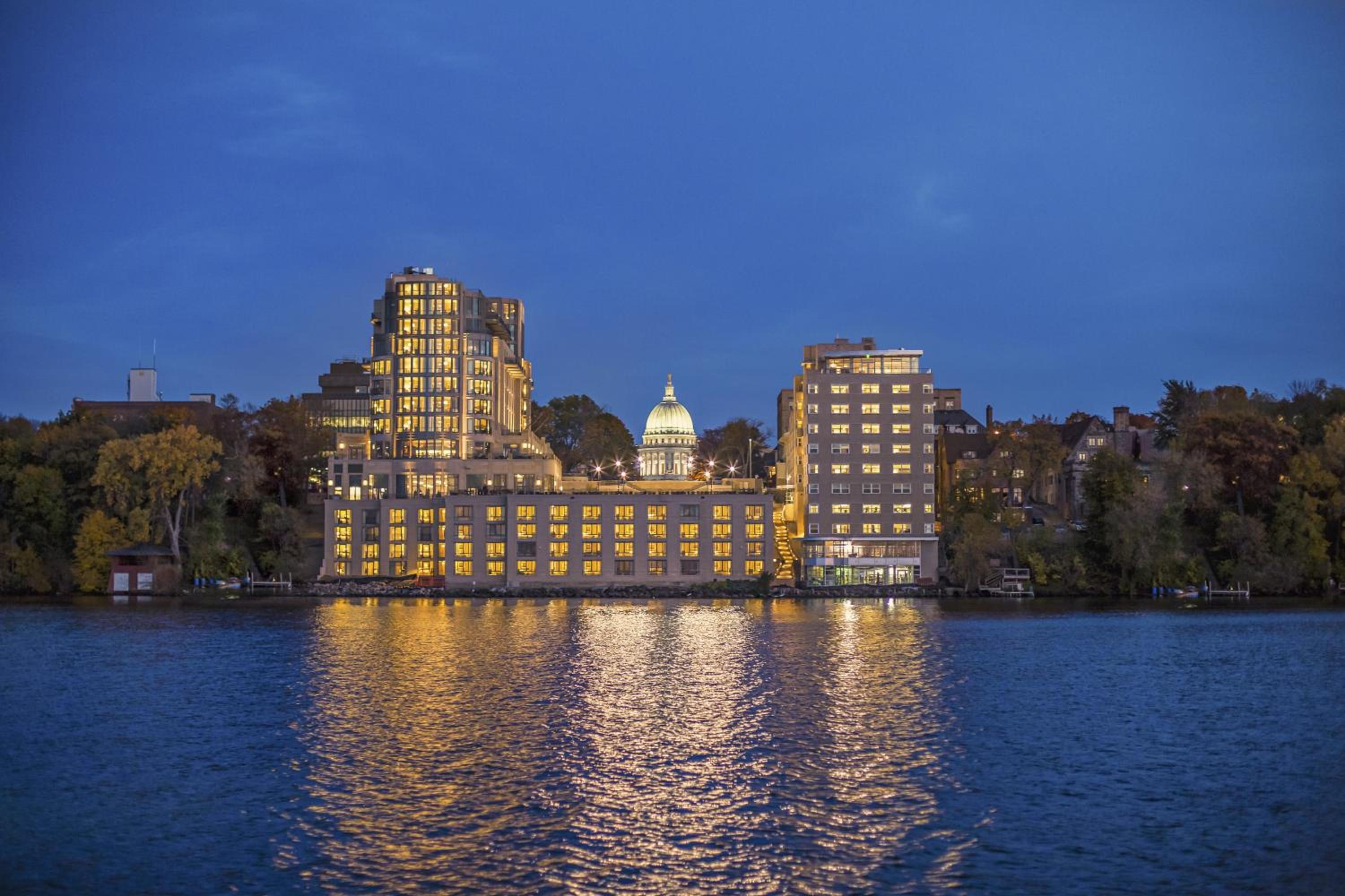 Waterfront buildings and an illuminated capitol dome reflected across dark rippling water at twilight.